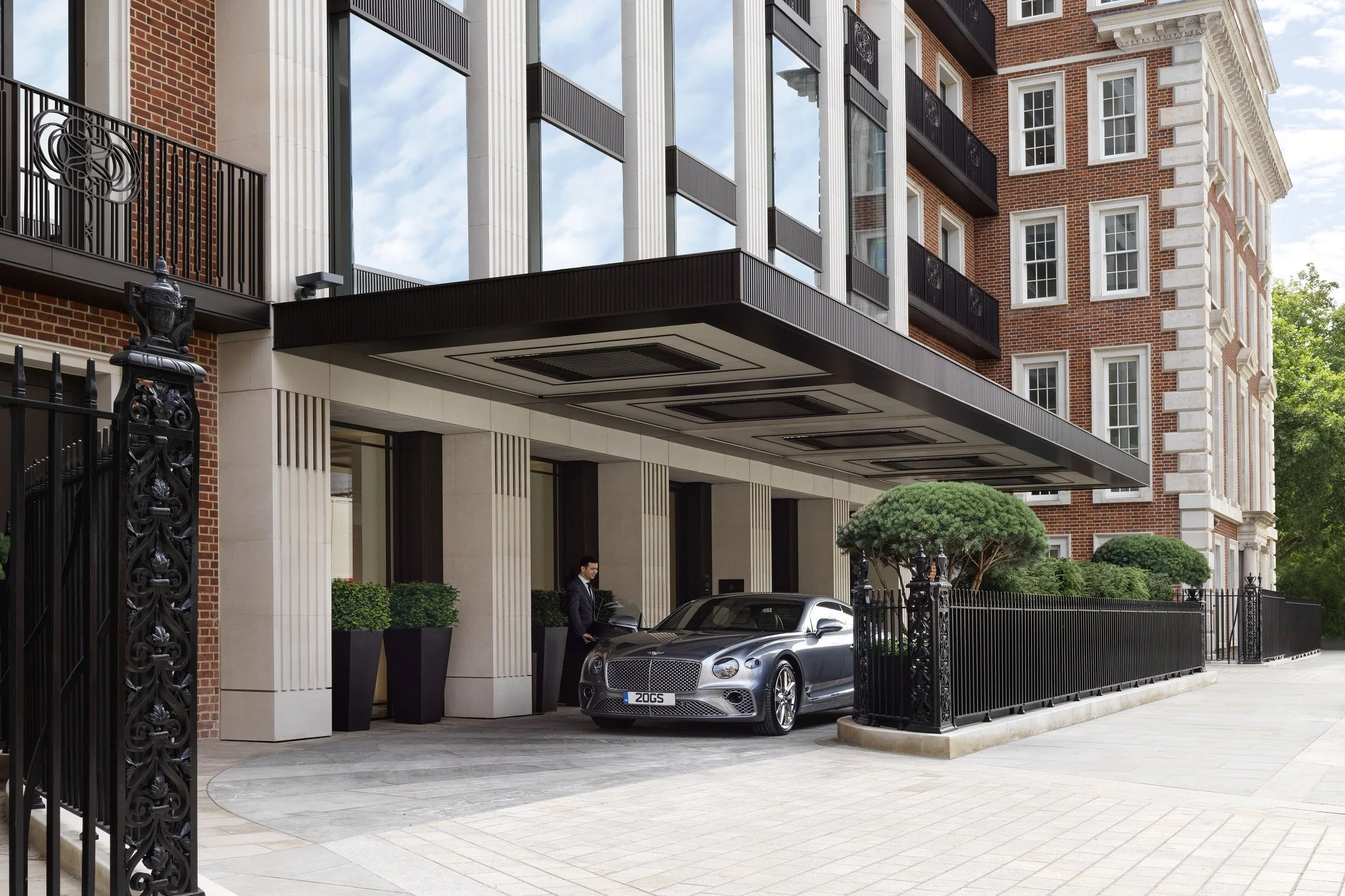 Luxury building entrance with modern architecture, black fence, lush green trees, a silver luxury car parked near the entrance, and a man in a suit standing beside the car.