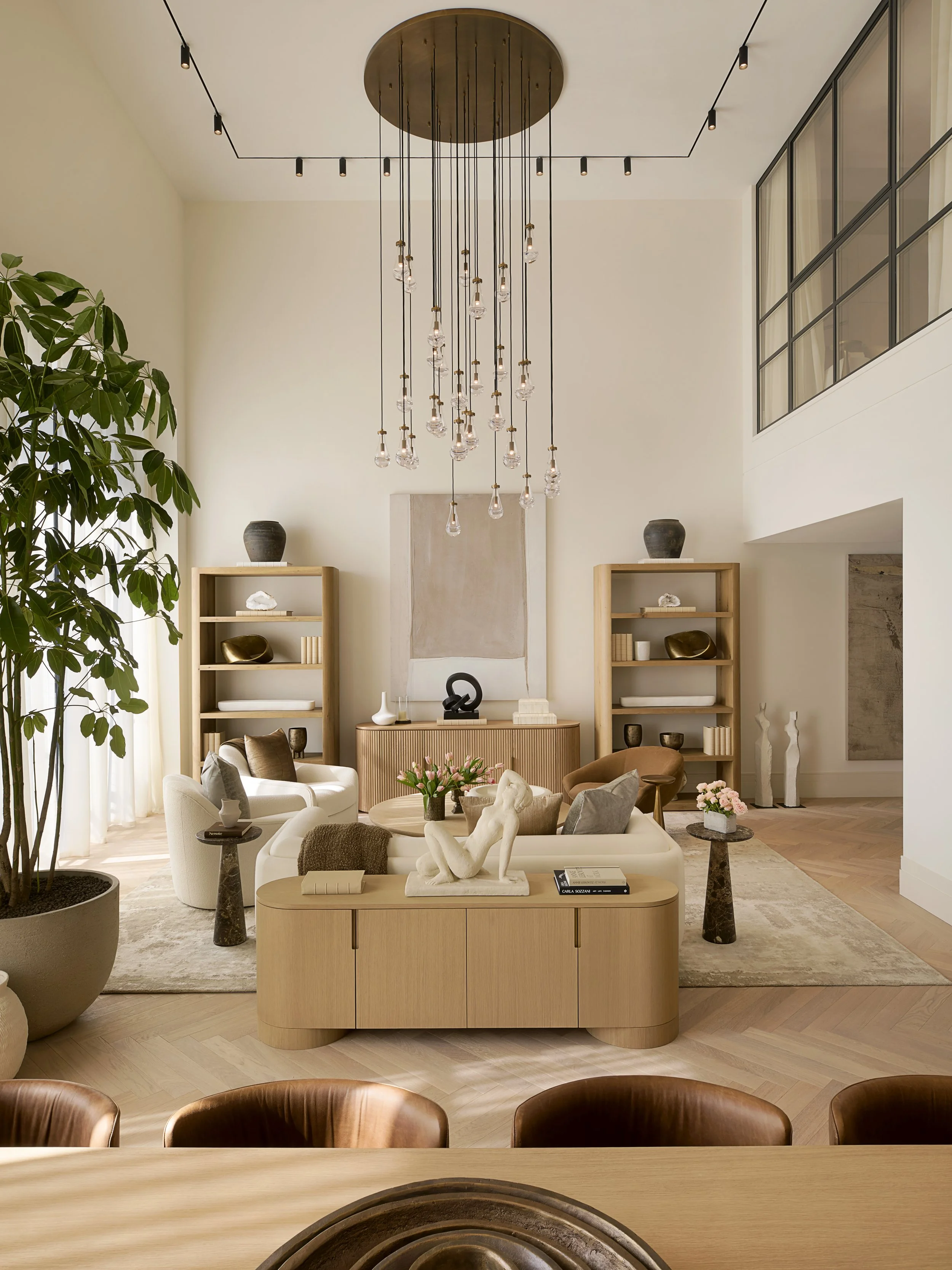 Modern living room with high ceiling, chandelier, beige sofa, armchairs, wooden bookshelves, and potted plants.