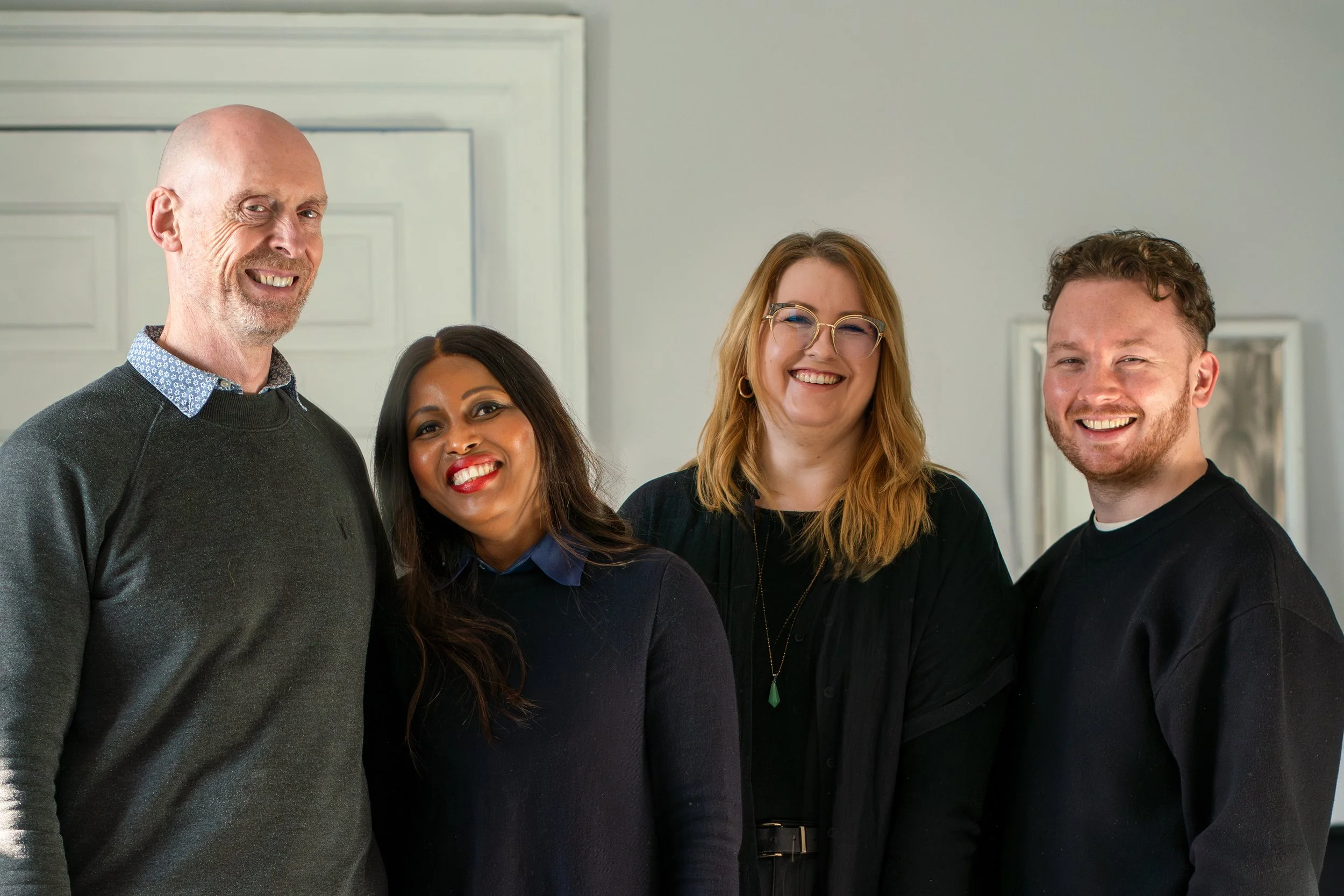 Group of four people smiling, standing indoors, diverse ages and ethnicities, casual attire.