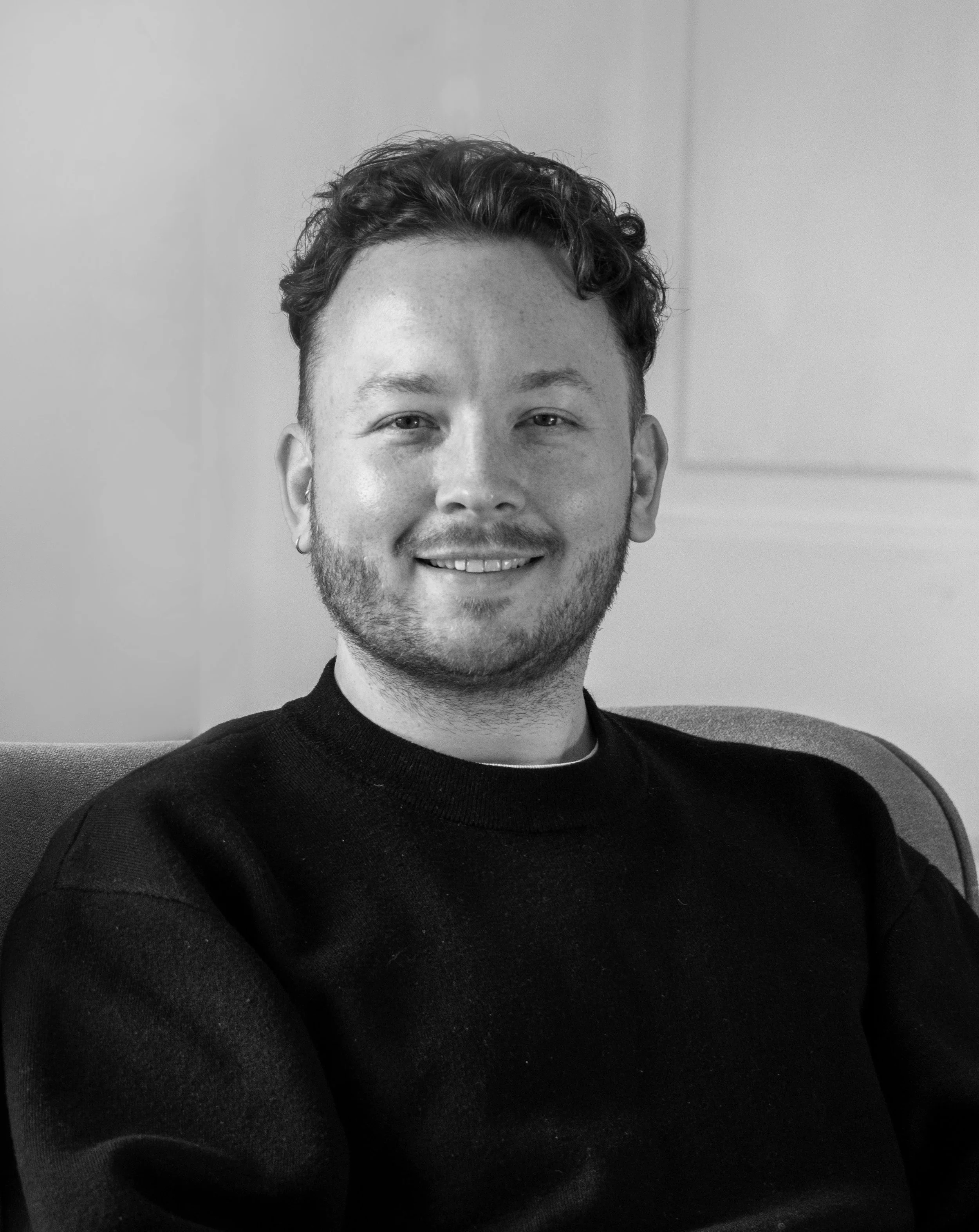 Black and white portrait of a smiling man with short curly hair, wearing a black sweater, sitting on a chair in front of a plain wall.