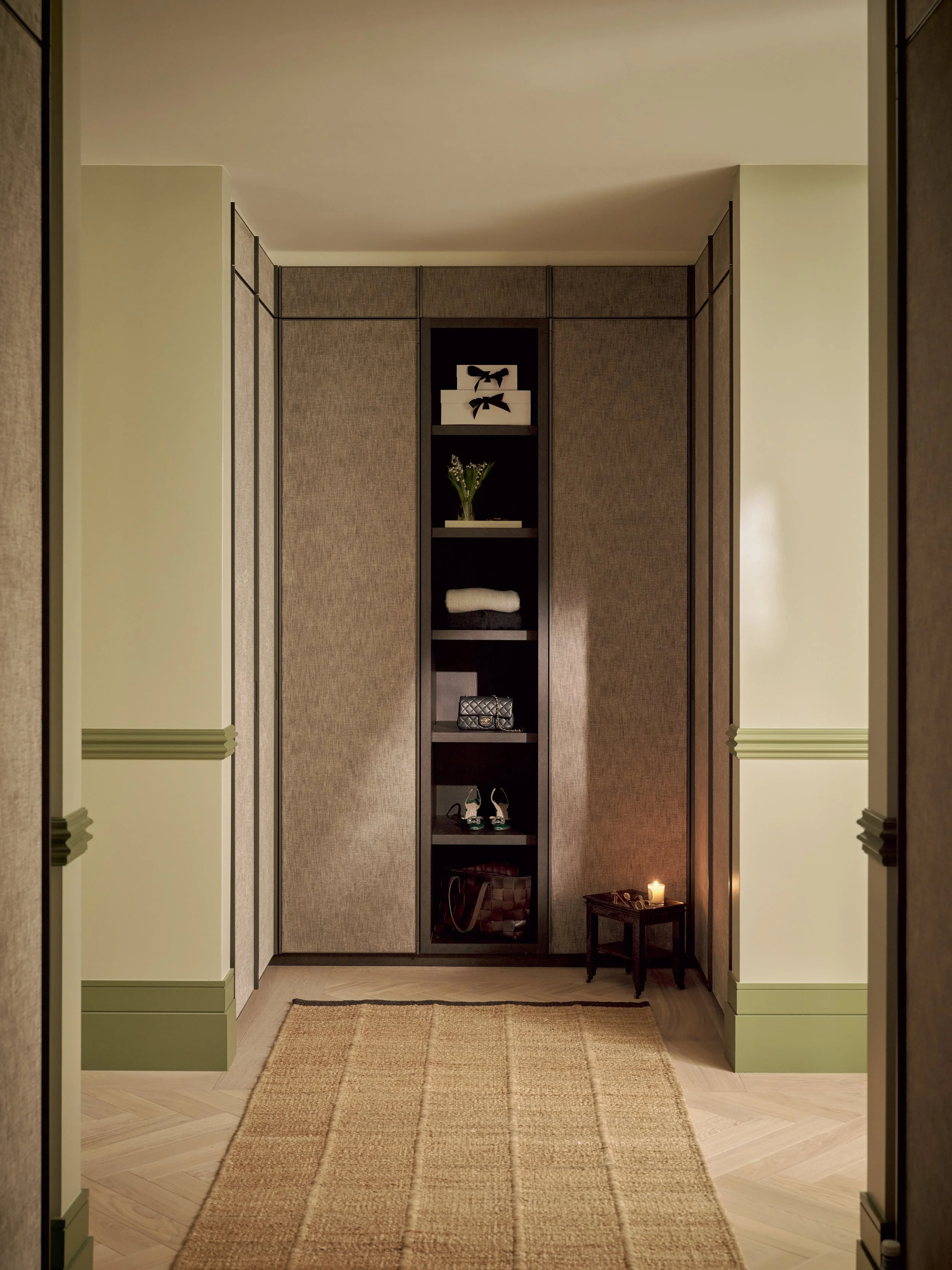 A narrow built-in shelf with various decorative items, a small side table with a lit candle, a beige rug, and tan wall panels in an interior hallway.