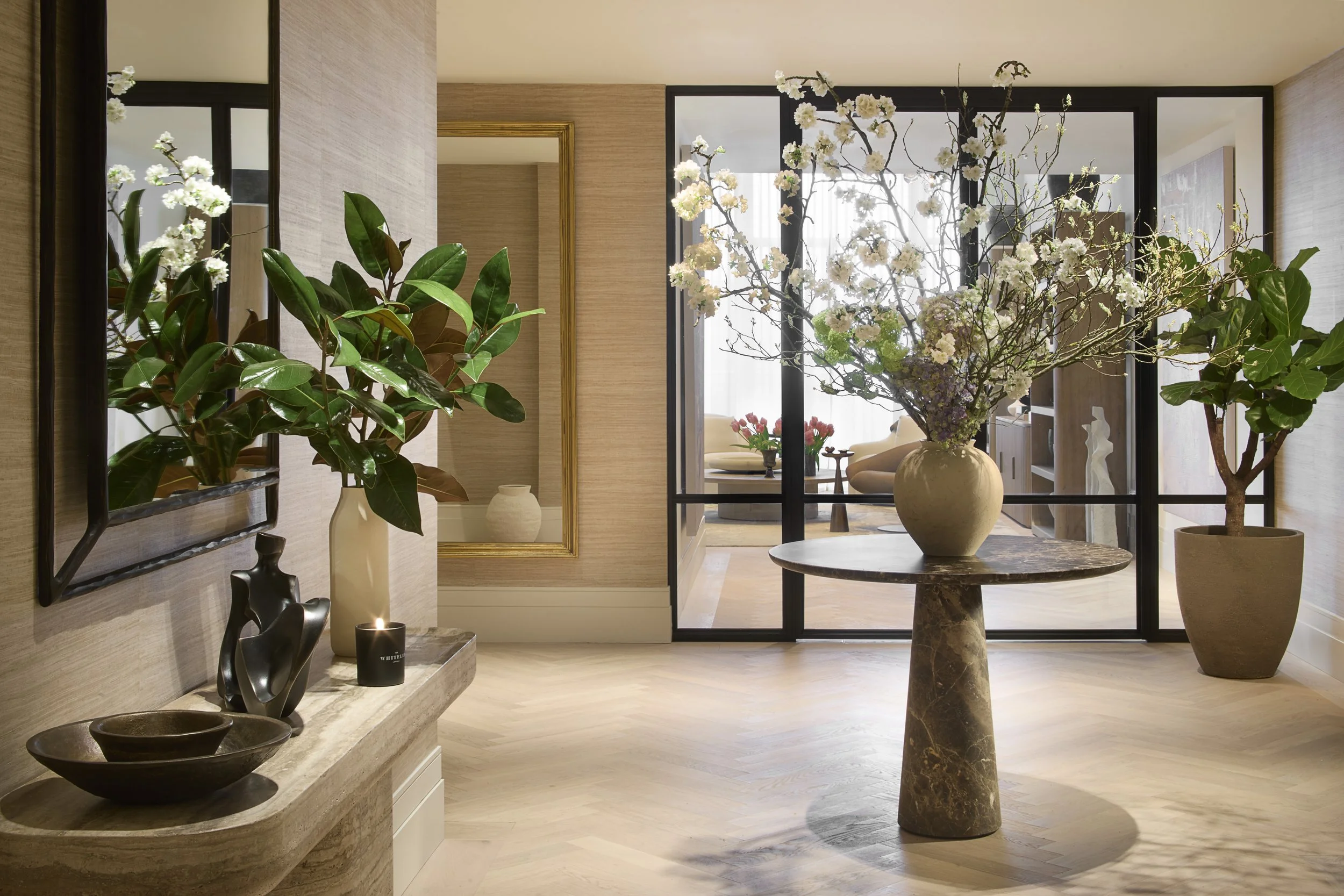 Elegant interior with a large floral arrangement in a prominent vase on a round marble table, potted plants, a mirror, and a view of the living room through a glass partition.