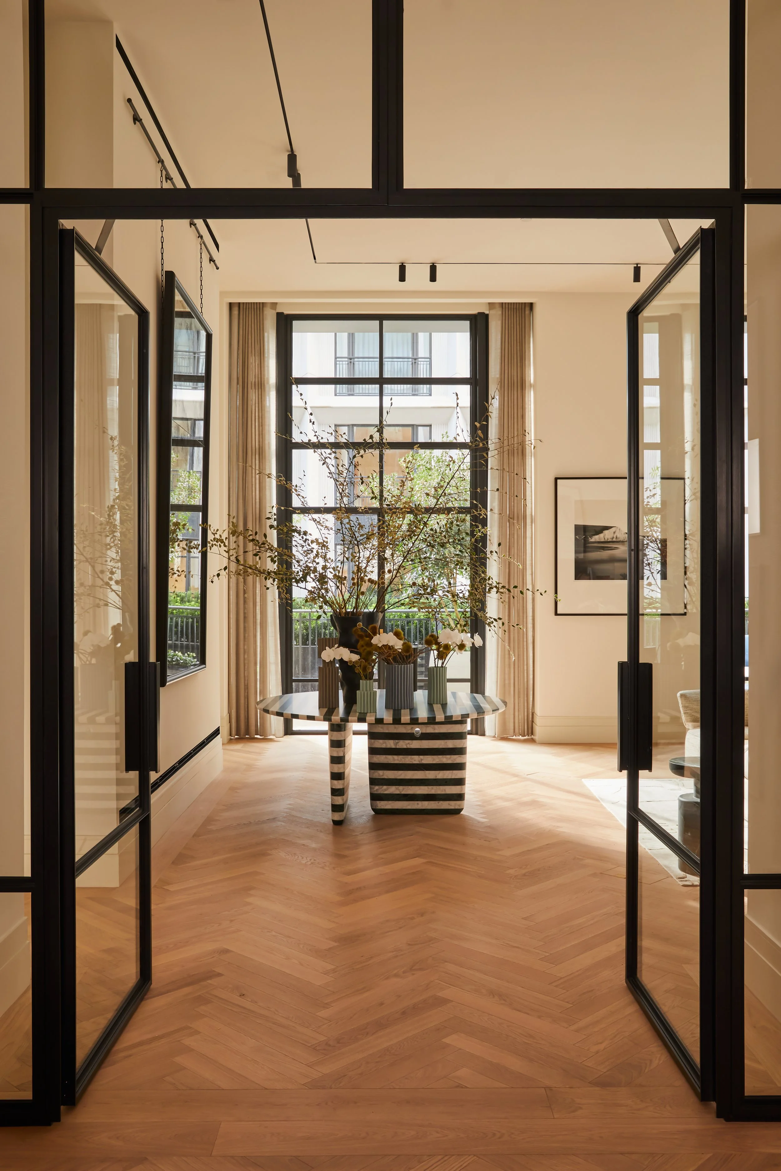 Modern interior room with large window, wooden floor, black-framed glass doors, and a striped black-and-white striped table with a large flower arrangement.