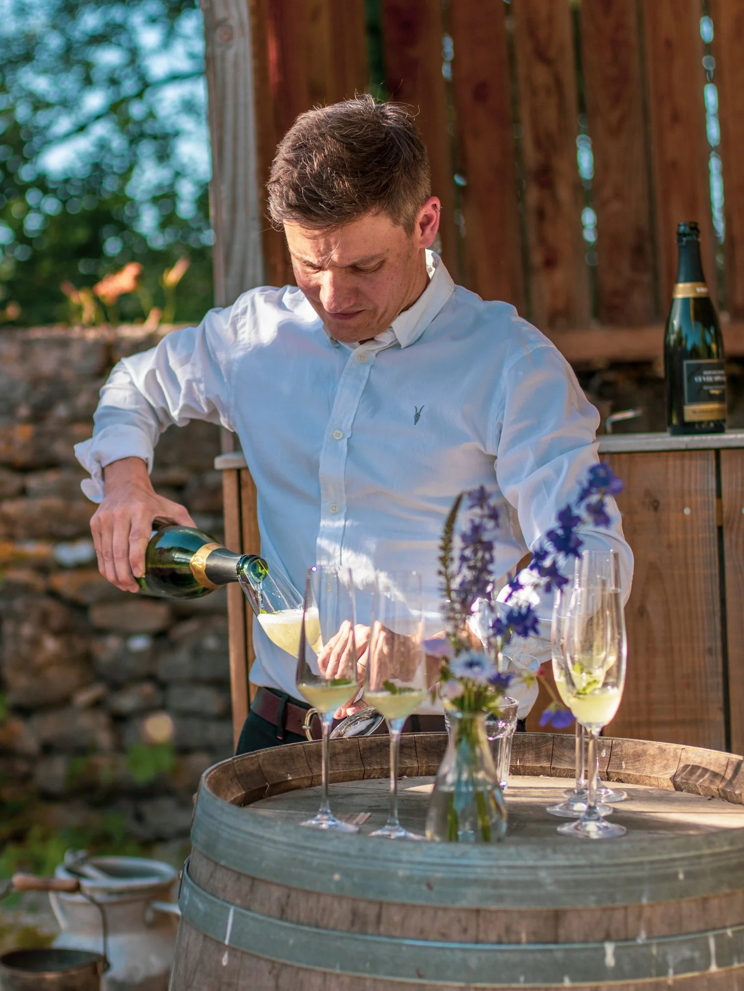 A man in a white shirt pours champagne into glasses on a table outdoors, with a wooden fence and greenery in the background.