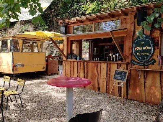 A rustic outdoor food stall made of wood with a serving window, surrounded by outdoor seating including a red table and chairs, and a yellow food truck with a yellow umbrella nearby. Green foliage is visible above.