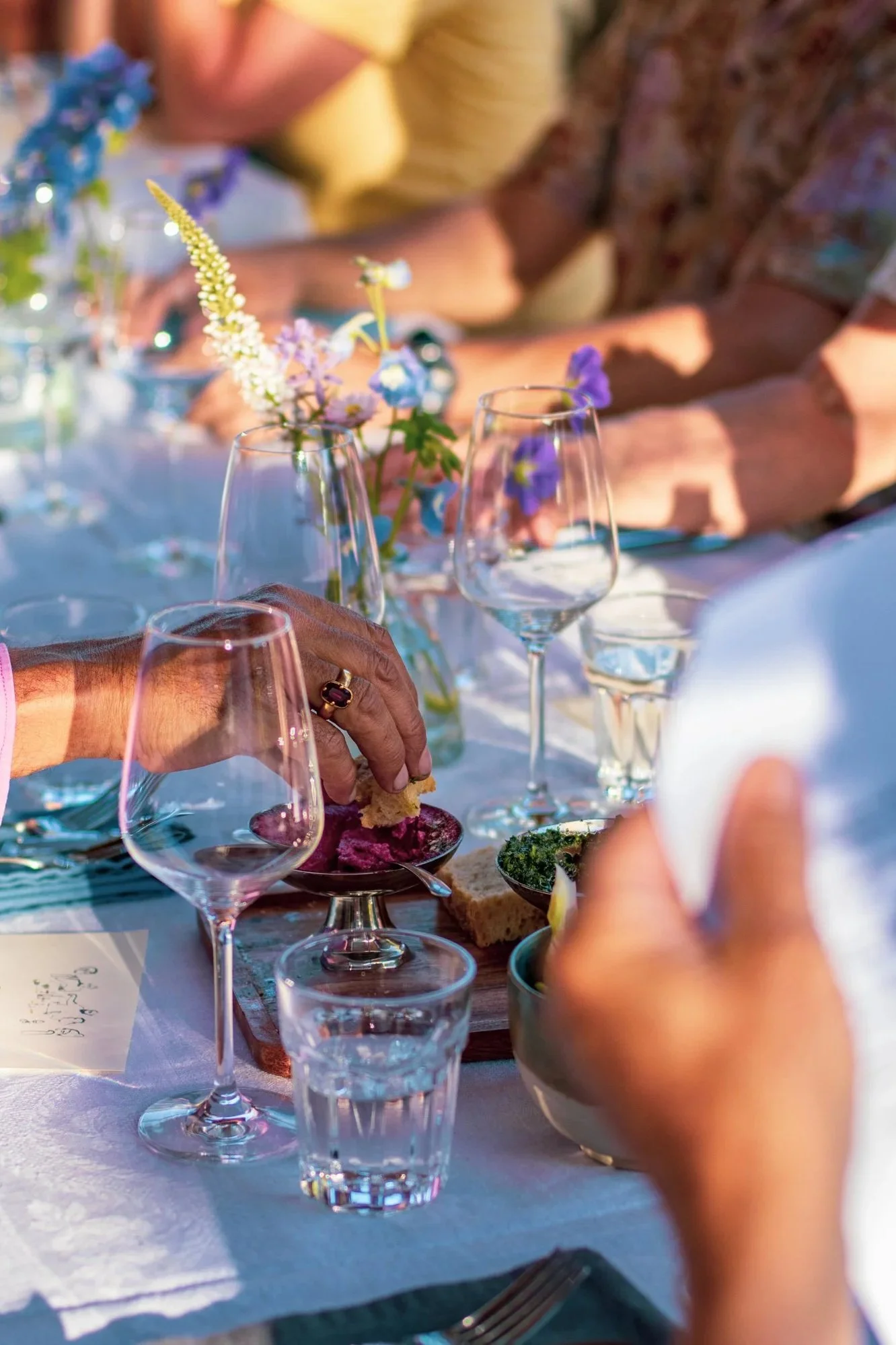 A close-up of a table set for a meal outdoors, featuring several wine glasses, a small glass of water, a dish with food, and a floral centerpiece. People are gathered around, with their hands reaching for food or drinks.