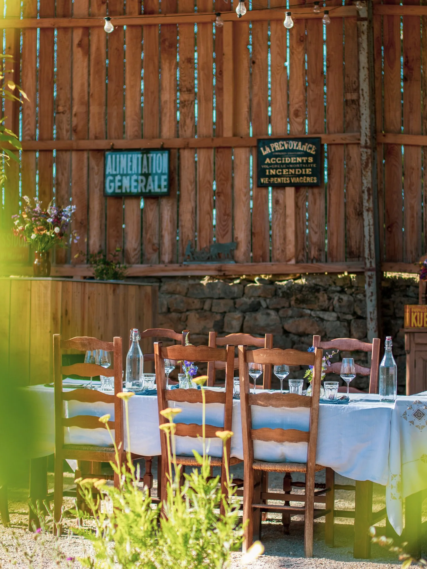 A round dining table set outdoors with a white tablecloth, wooden chairs, wine glasses, and bottles, surrounded by a wooden fence with garden signs and flowers.