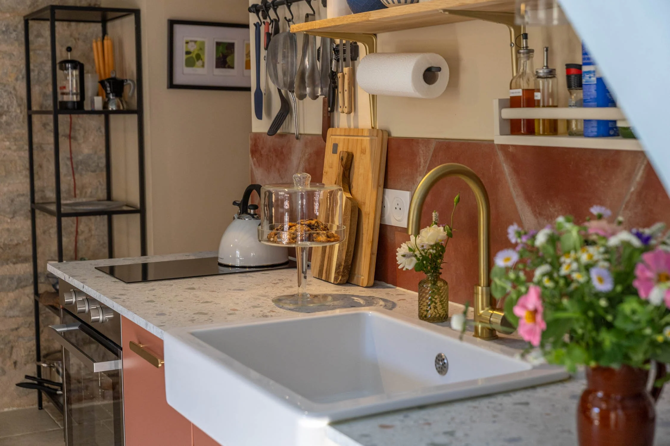 A kitchen countertop with a white farmhouse sink, a vase of flowers, a glass cake stand with cookies, a wooden cutting board, various knives, and paper towels. In the background, there is a spice rack, framed artwork, and a shelf with a coffee maker, candles, and other items.