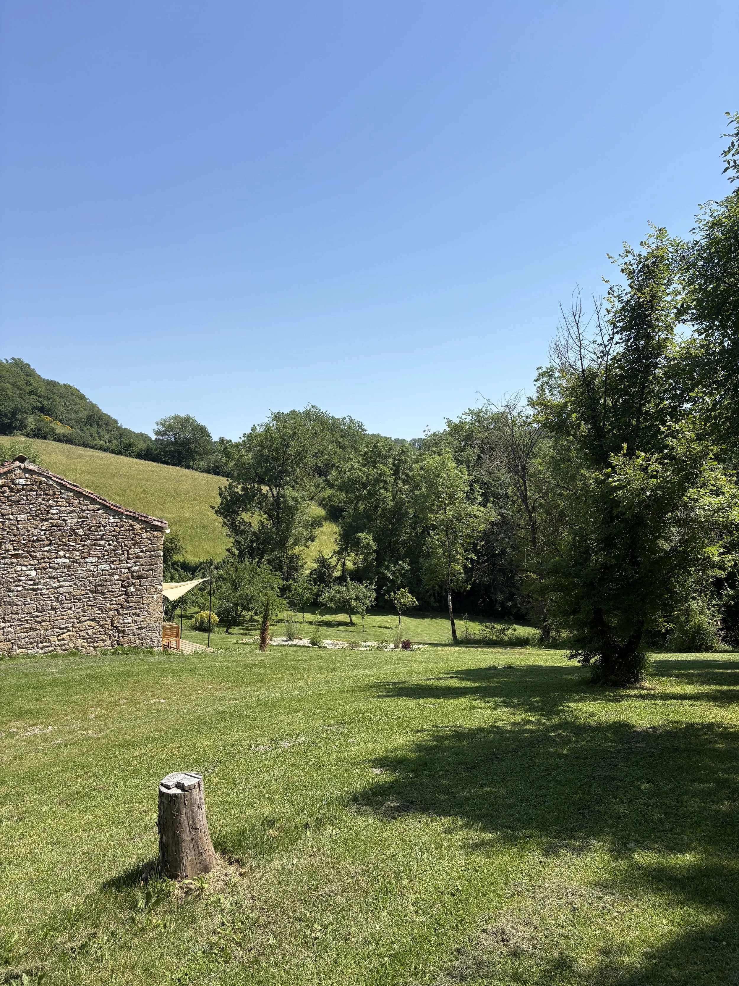 A scenic outdoor landscape featuring a grassy yard with a stone building to the left, surrounded by trees and a lush green hill in the background under a clear blue sky.