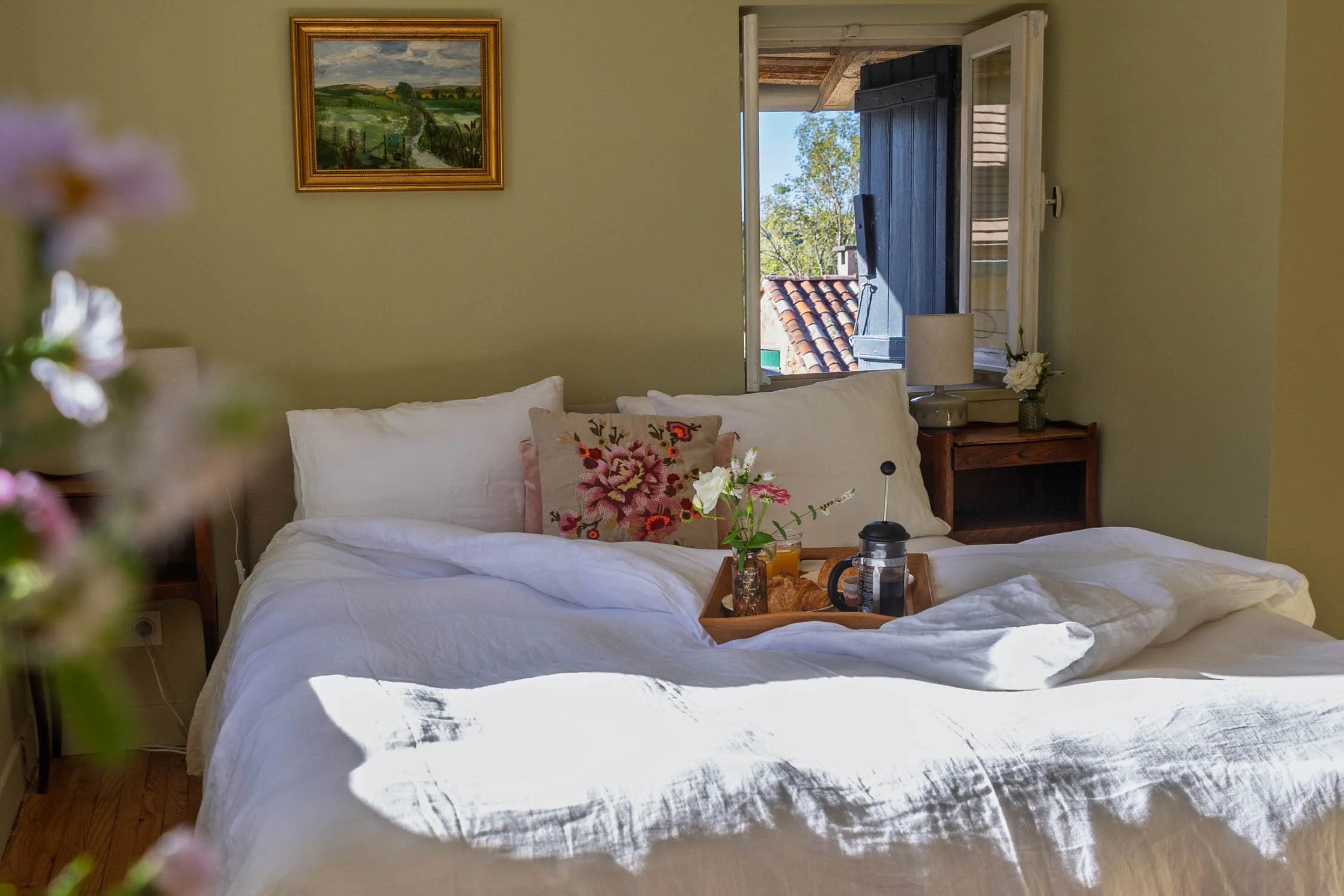 A bedroom with an unmade bed, white linens, and a floral pillow. On the bed, there is a tray with a glass of orange juice, a croissant, and a French press. The room has a window with an open shutter, showing rooftops and trees outside. A picture of a landscape hangs on the wall, and a small nightstand with a lamp and flowers sits next to the bed.