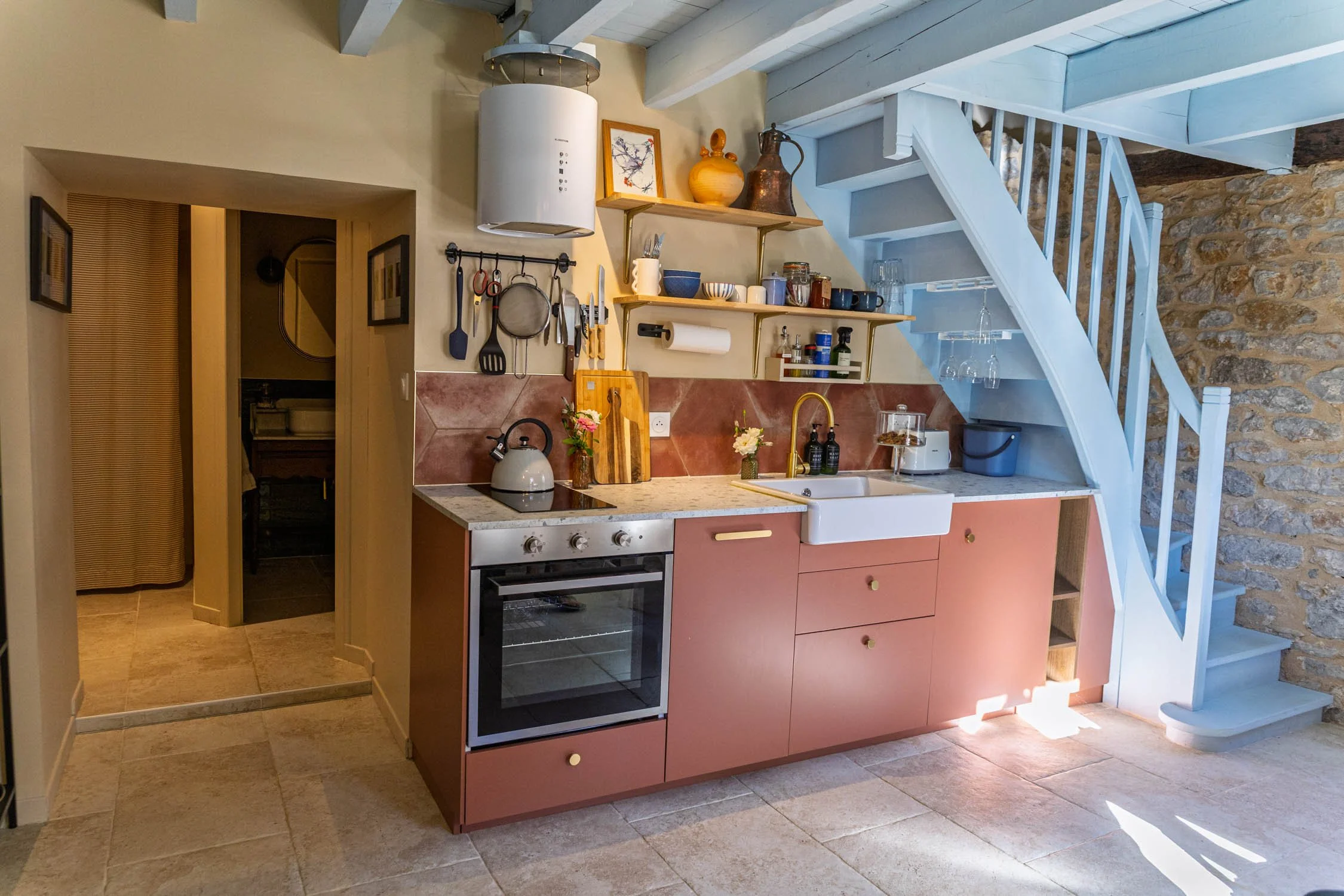 A cozy kitchen corner with pink cabinets, a white farmhouse sink, a stovetop, and wooden open shelves holding dishes, bowls, and decorative vases. A white range hood hangs above, and a stone wall is visible on the right side. The space features wooden beams and light-colored tiled flooring.