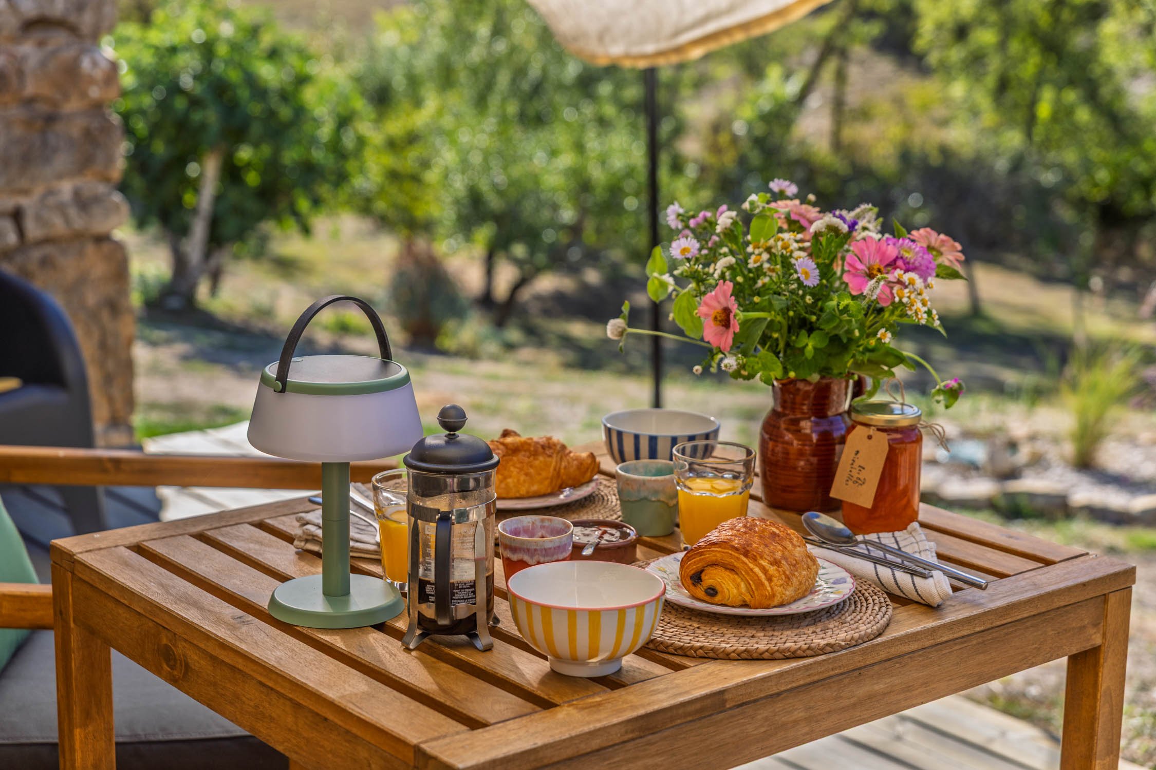 An outdoor breakfast table with croissants, bowls, cups, a French press, a lamp, a bouquet of pink, purple, and white flowers, glasses of orange juice, and a sunny garden in the background.