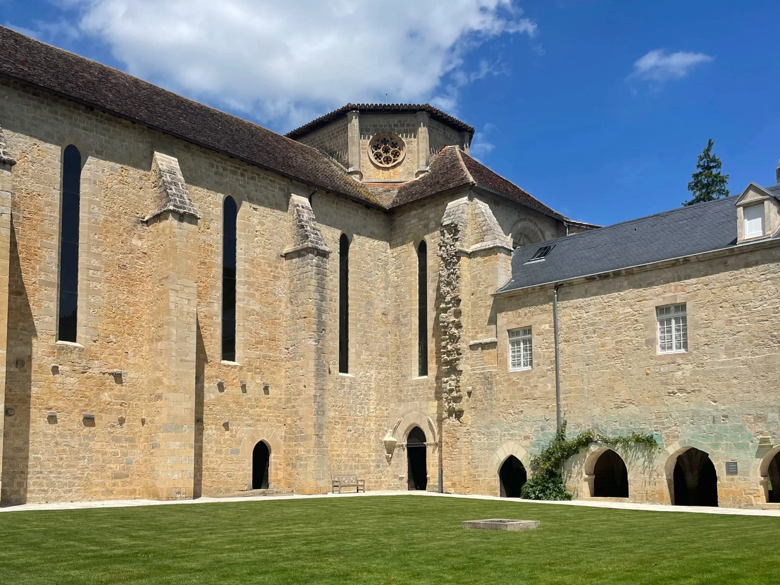 A stone church with gothic windows and a circular rose window, surrounded by a grassy courtyard, under a blue sky with clouds.