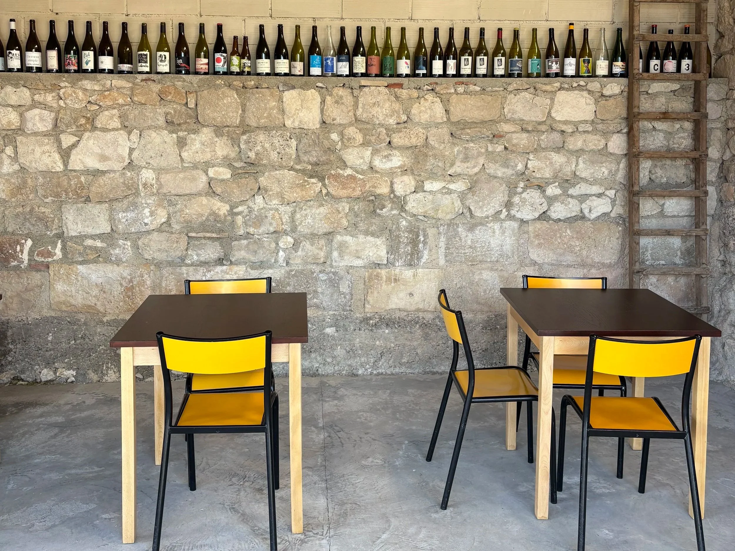 Interior of a rustic restaurant or cafe with a stone wall and shelves holding bottles with creative labels, four tables with black tops, and yellow chairs.