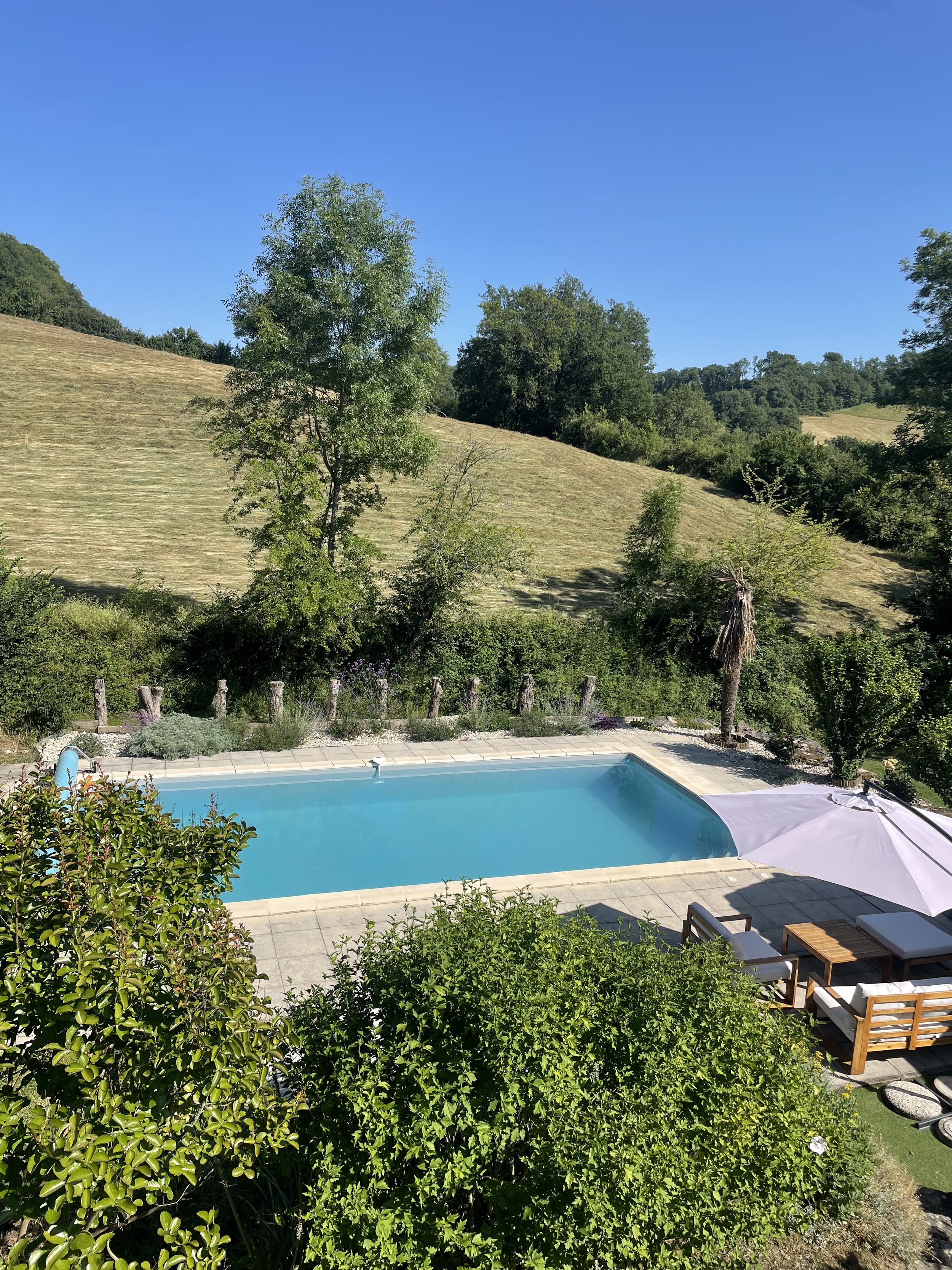 Swimming pool surrounded by greenery and rolling hills under clear blue sky.