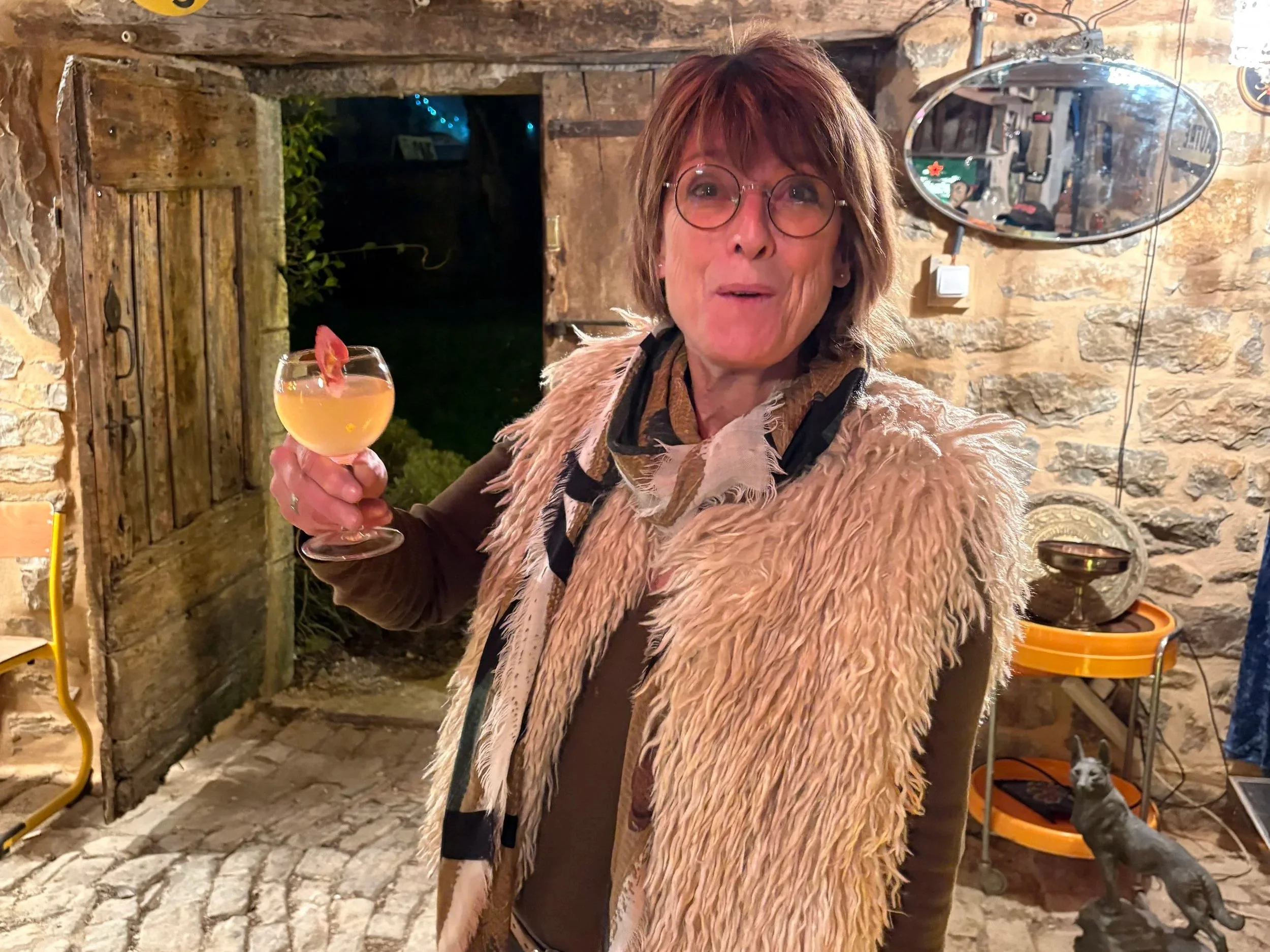 A woman with brown hair and glasses holding a cocktail glass with a pink flower garnish, smiling indoors with stone walls, a mirror, and a small dog in a pet bed in the background.