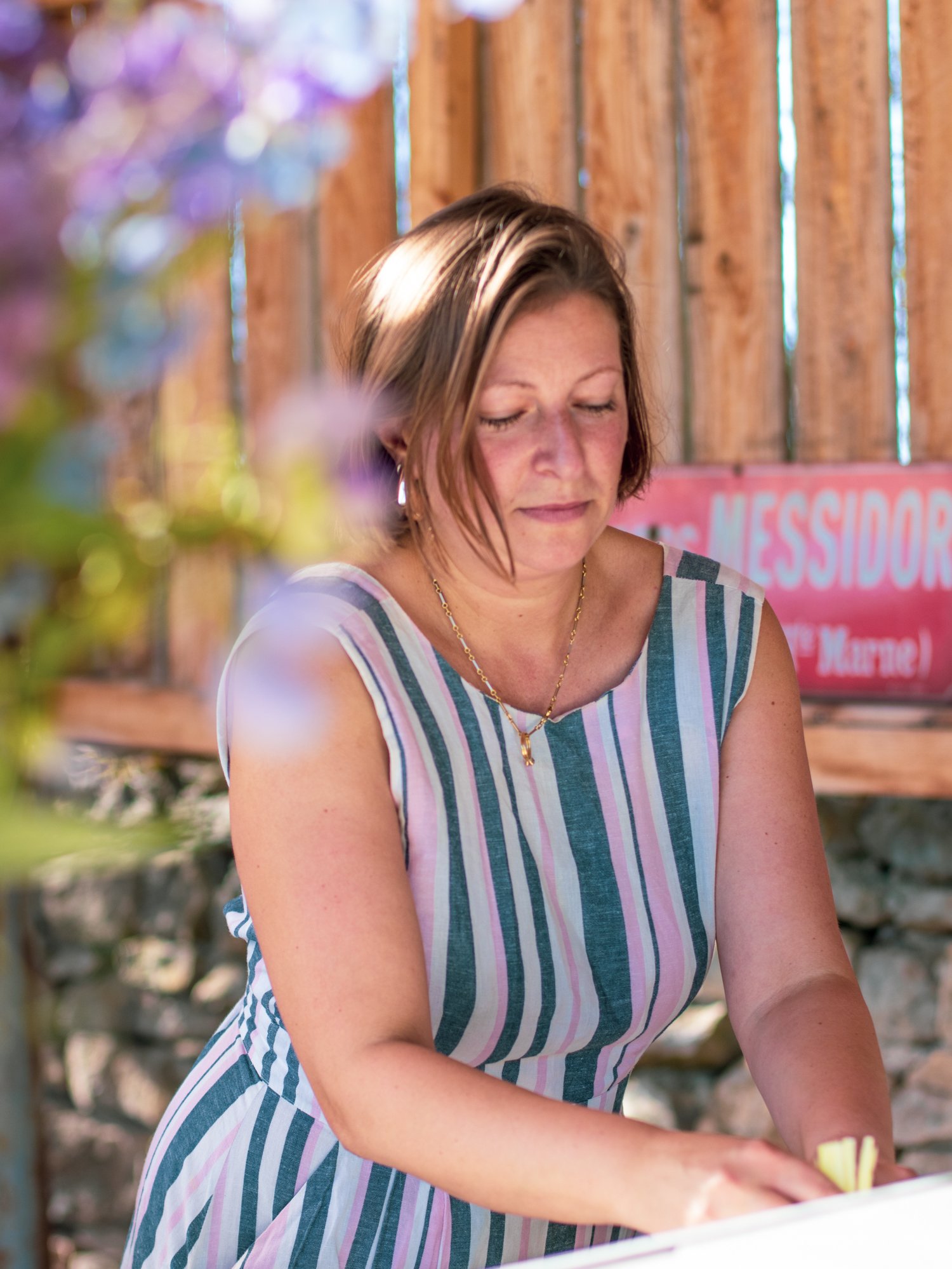 A woman with short brown hair and light skin, wearing a sleeveless striped dress, is looking down at the kitchen counter. She is outdoors near wooden fencing and some blurred colorful flowers.