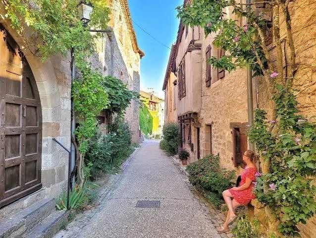 A narrow stone street with old buildings on both sides, greenery, and a girl in a red dress sitting on a stone wall.