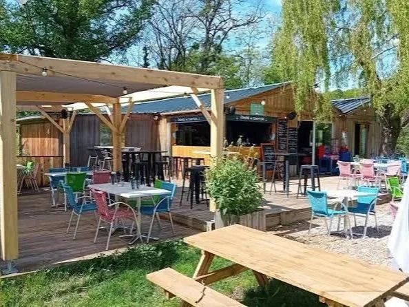 Outdoor patio with colorful chairs and tables, wooden deck, potted plant, and a small bar or food stand, surrounded by trees and a wooden fence.