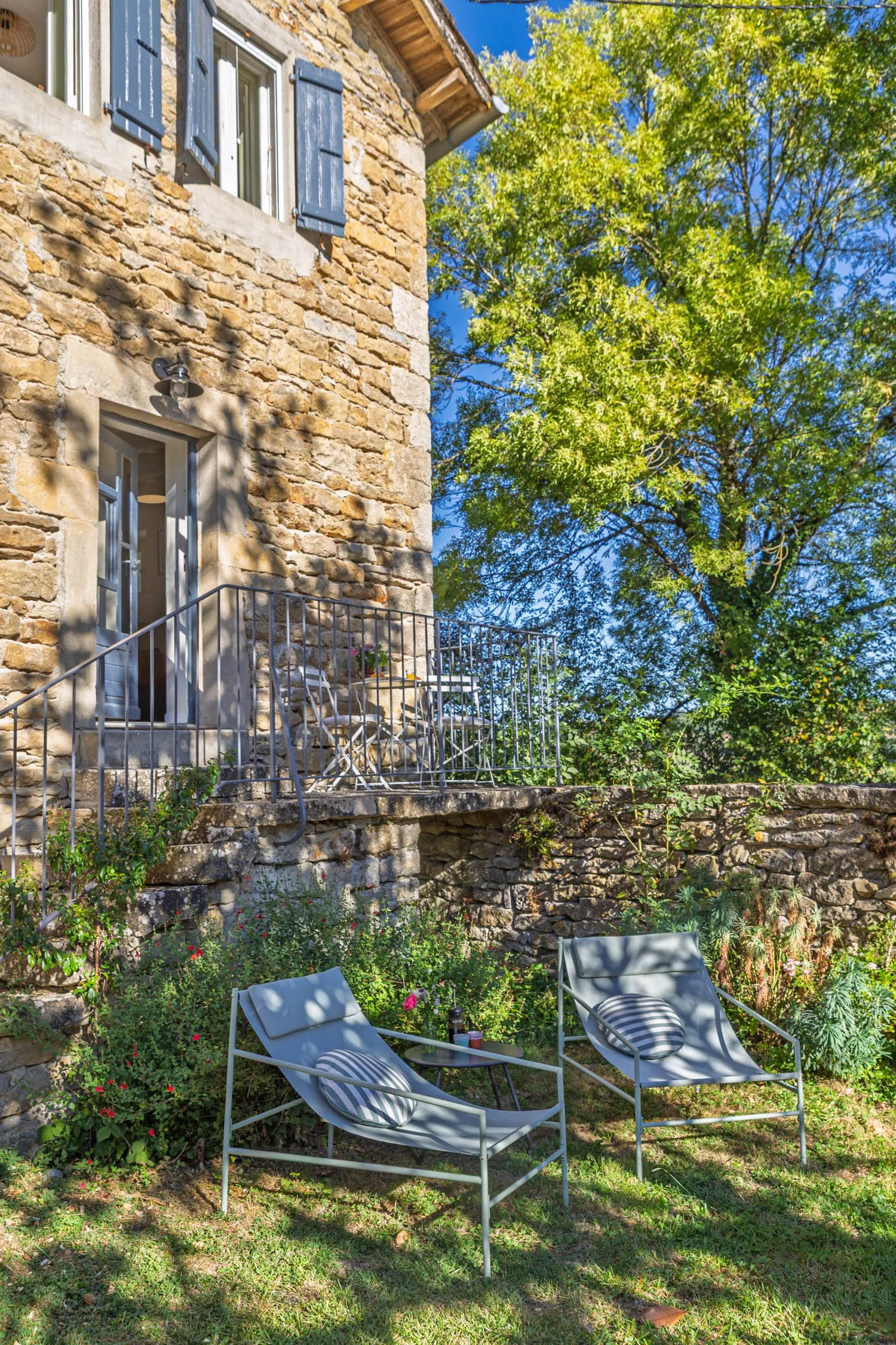 A cozy outdoor garden patio with two lounge chairs, a small table, lush green plants, and a stone house with blue shutters, set against a bright blue sky with a large leafy tree.