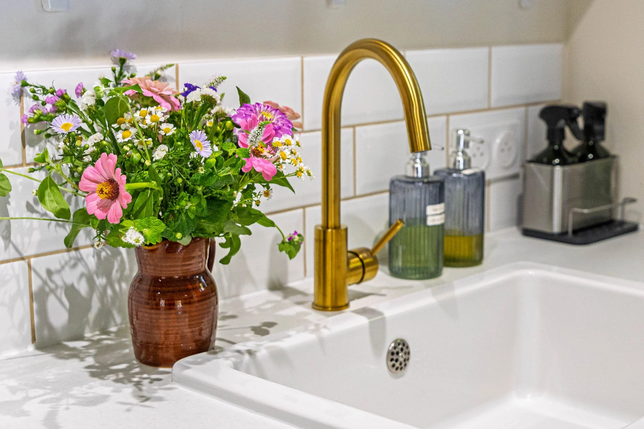 A kitchen sink area with a brown vase of mixed flowers, a gold faucet, two soap or sanitizer dispensers, and a coffee machine.