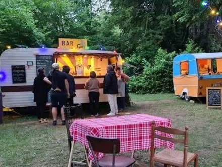 People gather at outdoor food trucks in a wooded area with string lights overhead and a table with a red checkered tablecloth in the foreground.