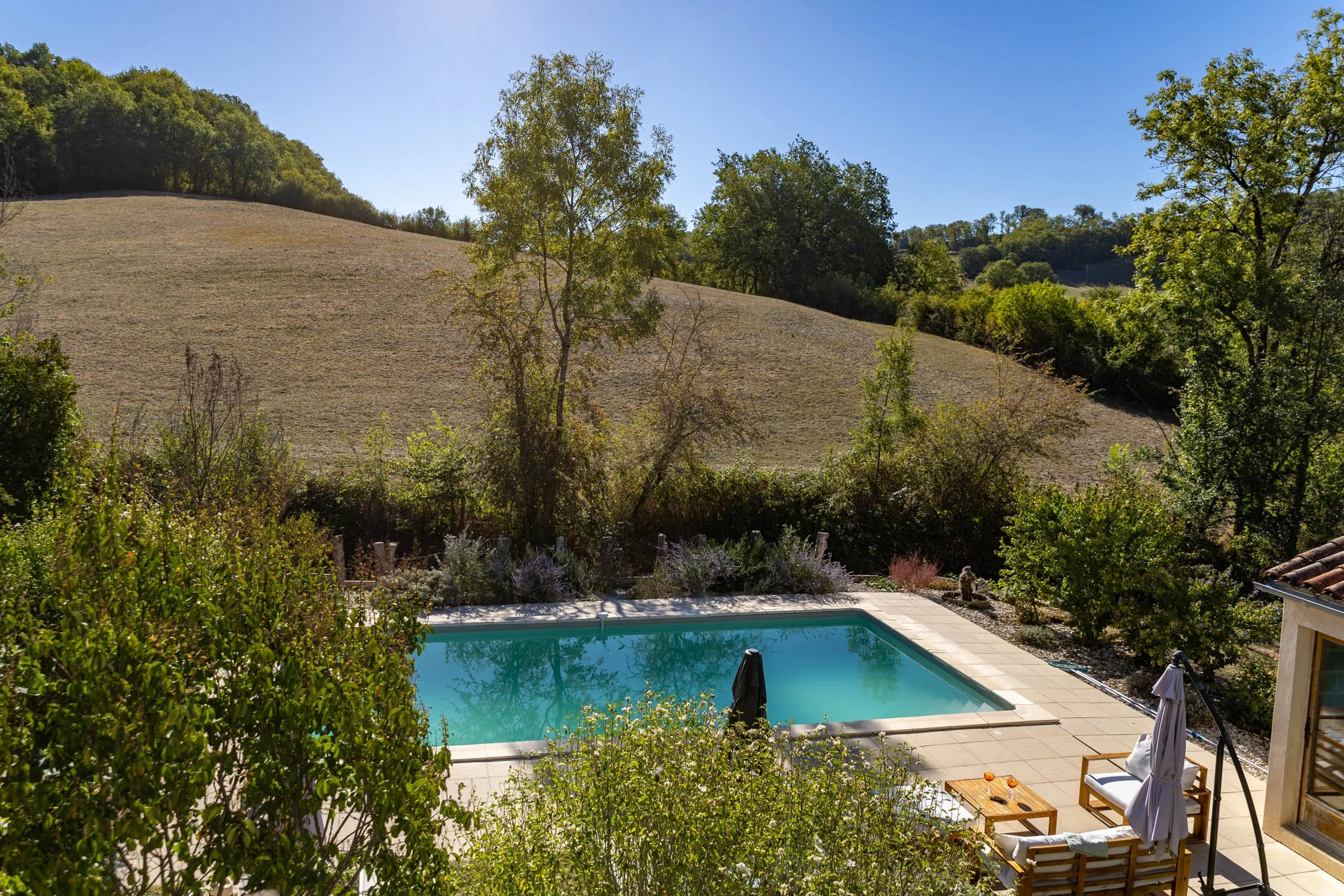 A backyard with a swimming pool, outdoor furniture, and lush greenery, overlooking a hillside with trees.