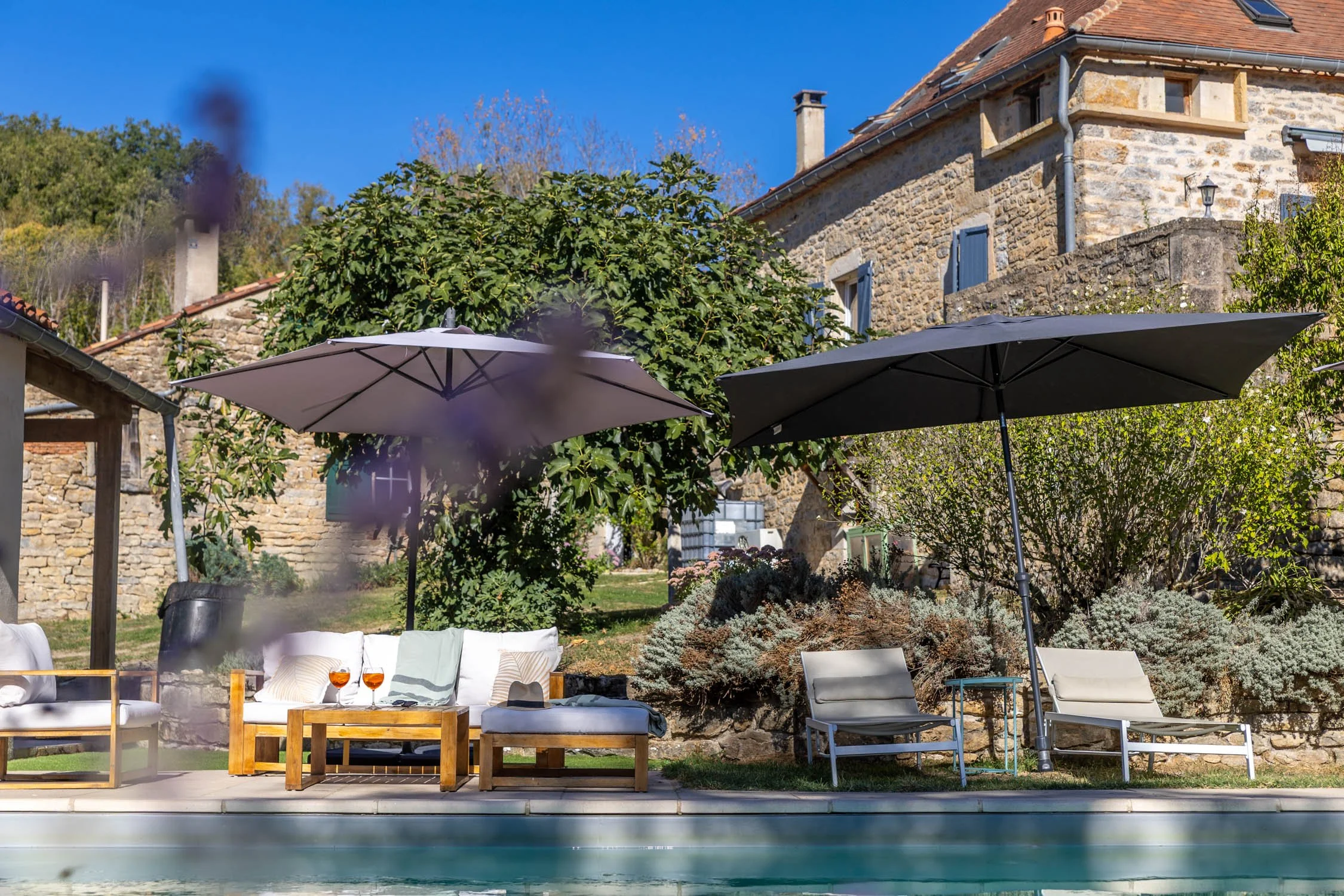 An outdoor patio area with lounge chairs, a sofa, a coffee table with two glasses of wine, large umbrellas, and a stone house in the background under a clear blue sky.