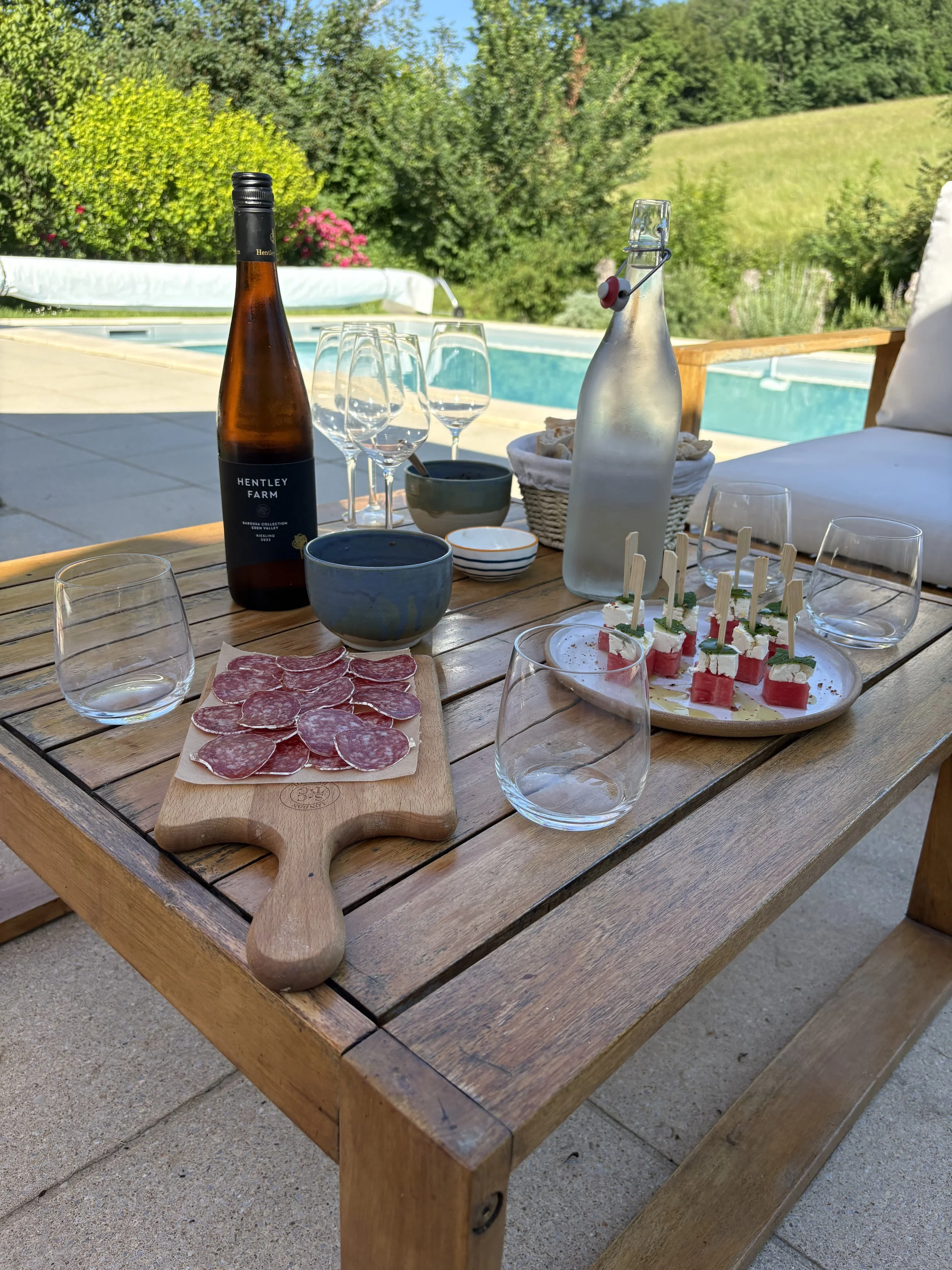 A wooden outdoor table set with wine glasses, a bottle of rose wine, a bottle of water, a small bowl, a charcuterie board with cured meats, and a cheese platter with small skewers, overlooking a backyard with a swimming pool and lush green trees and 