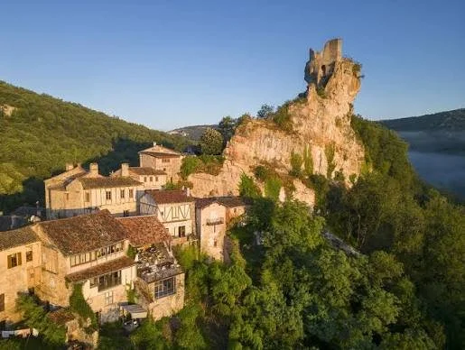 A cluster of old stone buildings with tiled roofs on a hillside, overlooking a river with a rocky cliff and ruins at the top surrounded by green trees.