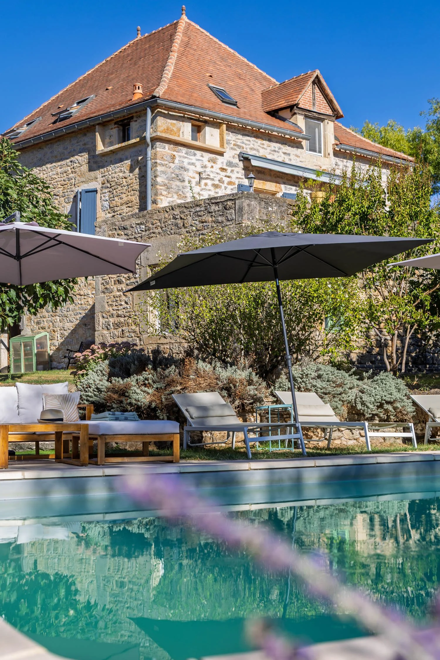 View of a stone house with a red tiled roof, set against a bright blue sky, with patio umbrellas, lounge chairs, a sofa, and pool in the foreground.