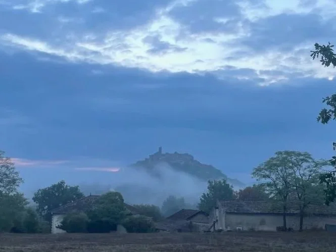A mountain with a castle on top, shrouded in fog, with a partly cloudy sky above and trees and rooftops in the foreground.