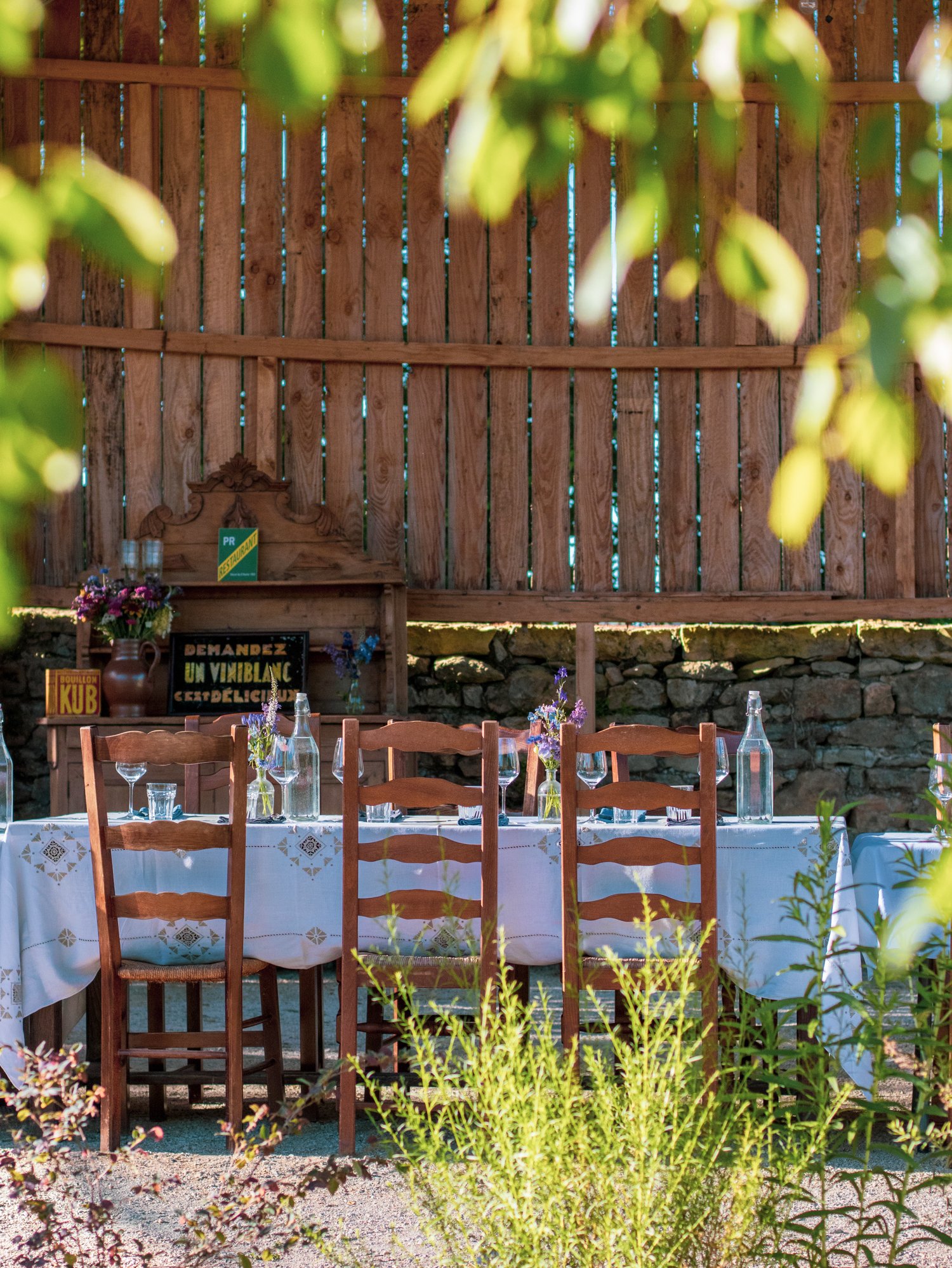 A table set for outdoor dining with flowers and glass bottles, in front of a wooden fence with greenery and plants in the foreground.