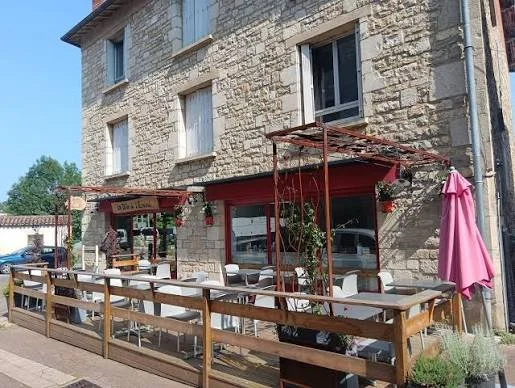 Outdoor seating area with tables and chairs outside a stone building, with a pink umbrella and potted plants.