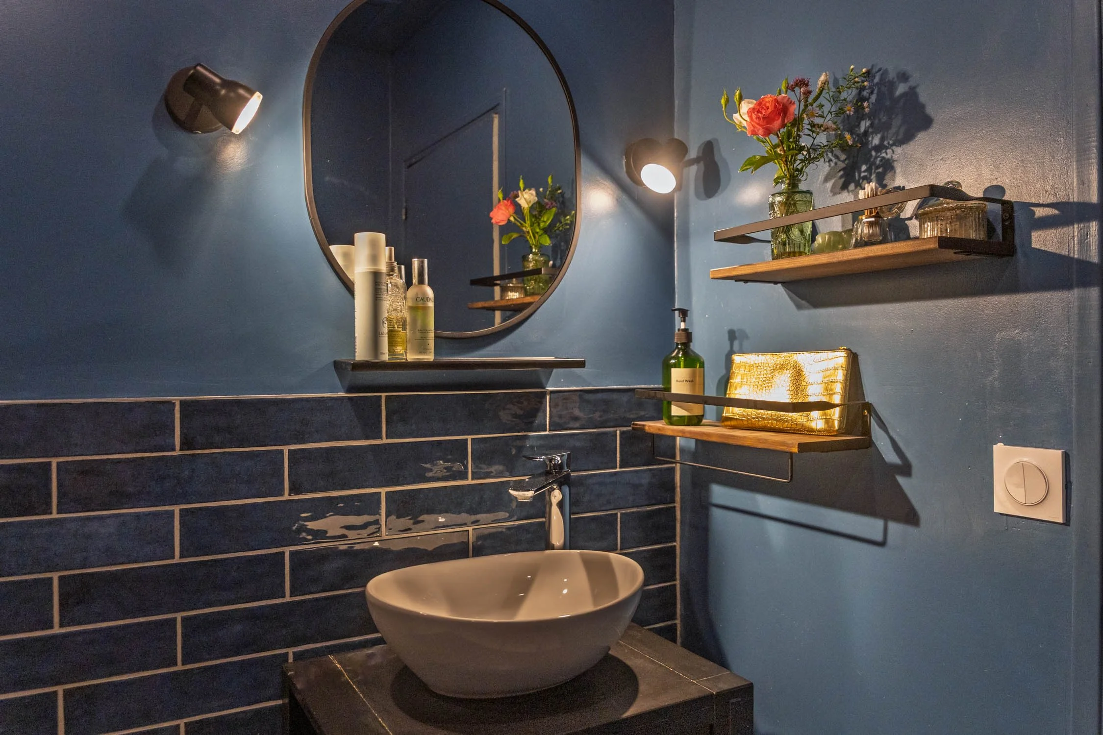Bathroom with a round mirror, white vessel sink, black tiled backsplash, wall-mounted black hardware, and blue painted walls decorated with white flowers and small green figurines.