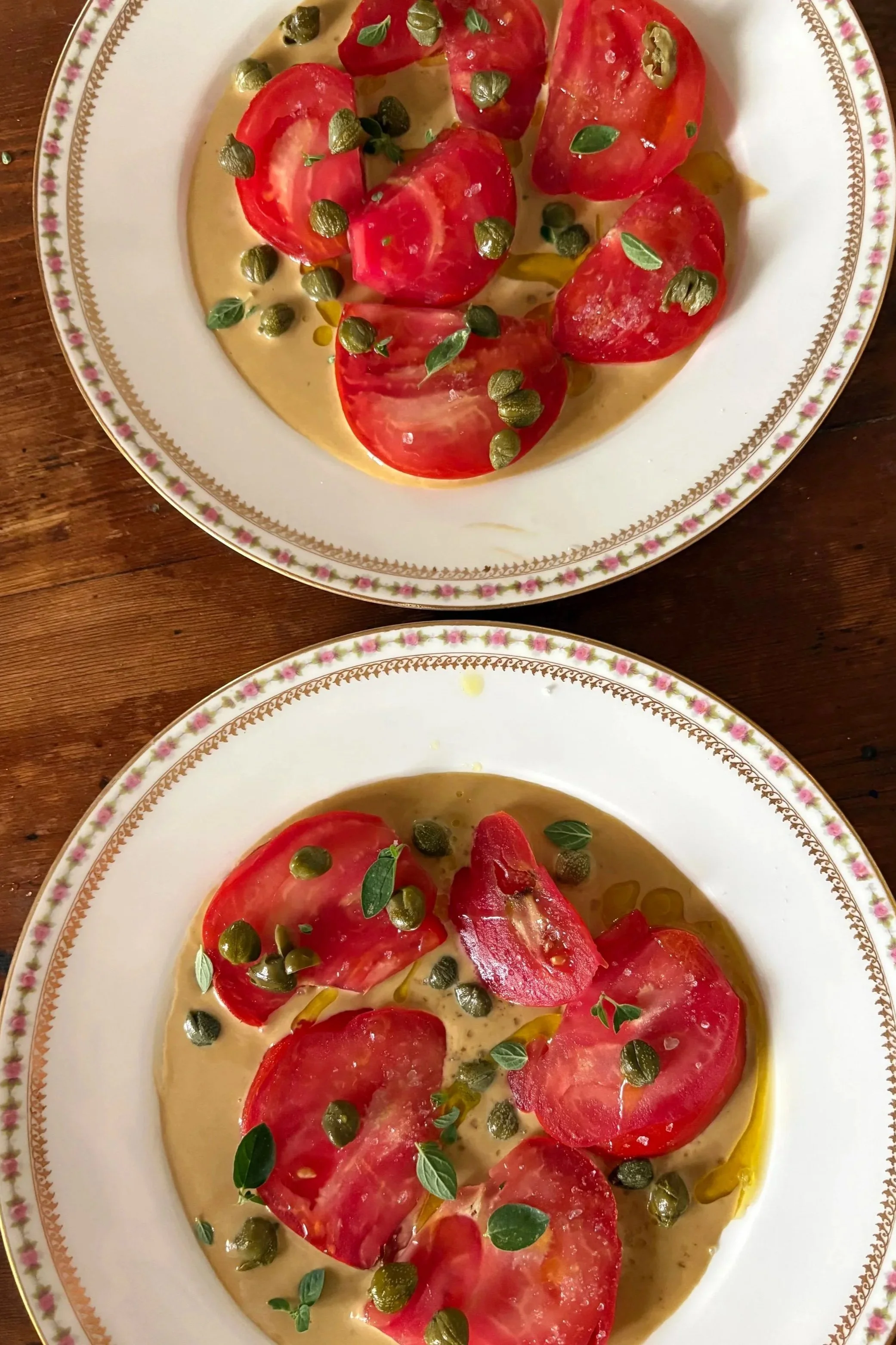 A white plate with a floral gold and pink trim border, holding sliced red tomatoes, small green herbs, capers, and a creamy yellow dressing or sauce.