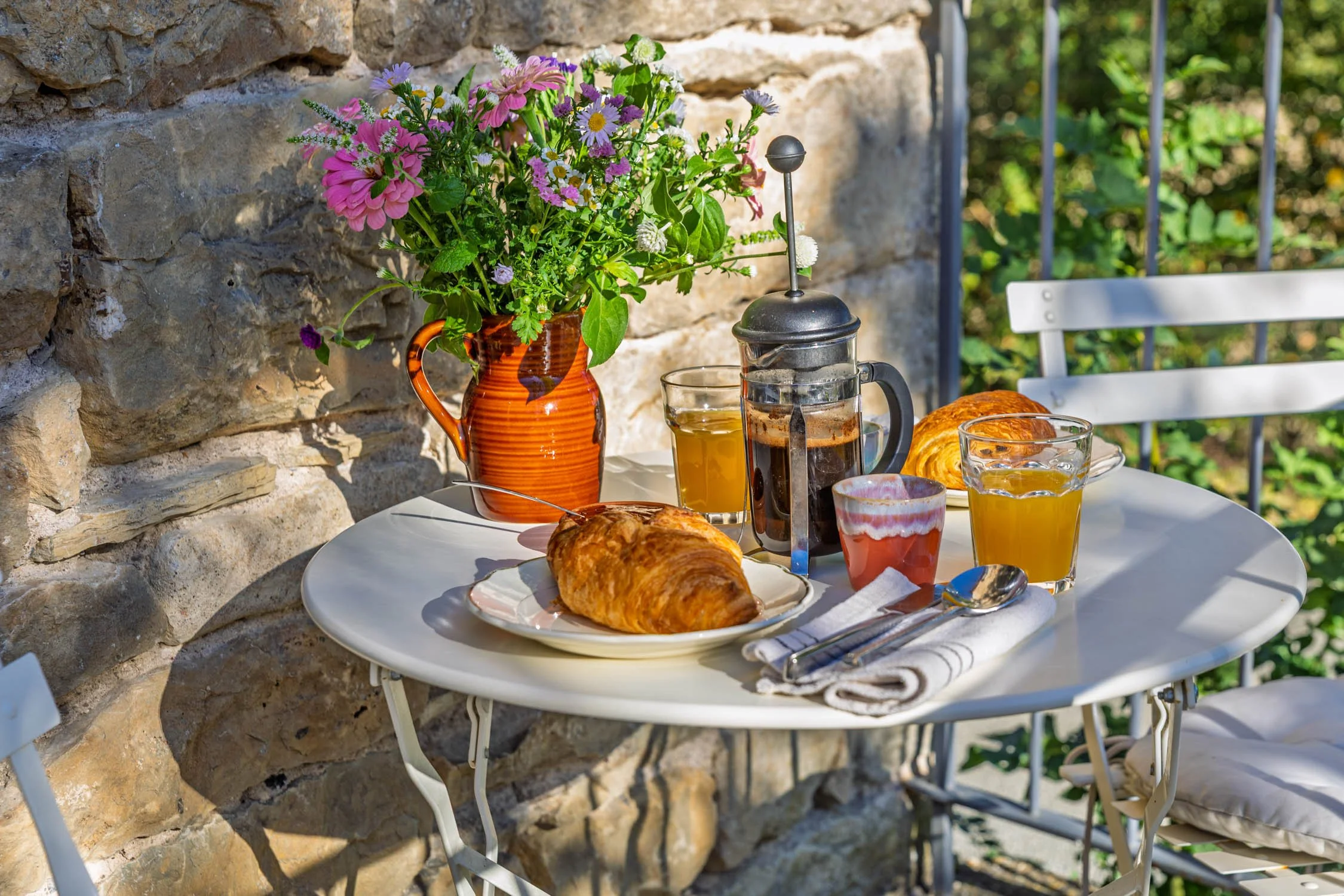 A round white table set for breakfast with croissants, glasses of juice, and coffee. There is a tall vase with purple and pink flowers and a French press with coffee. The table is outdoors against a stone wall with a garden in the background.