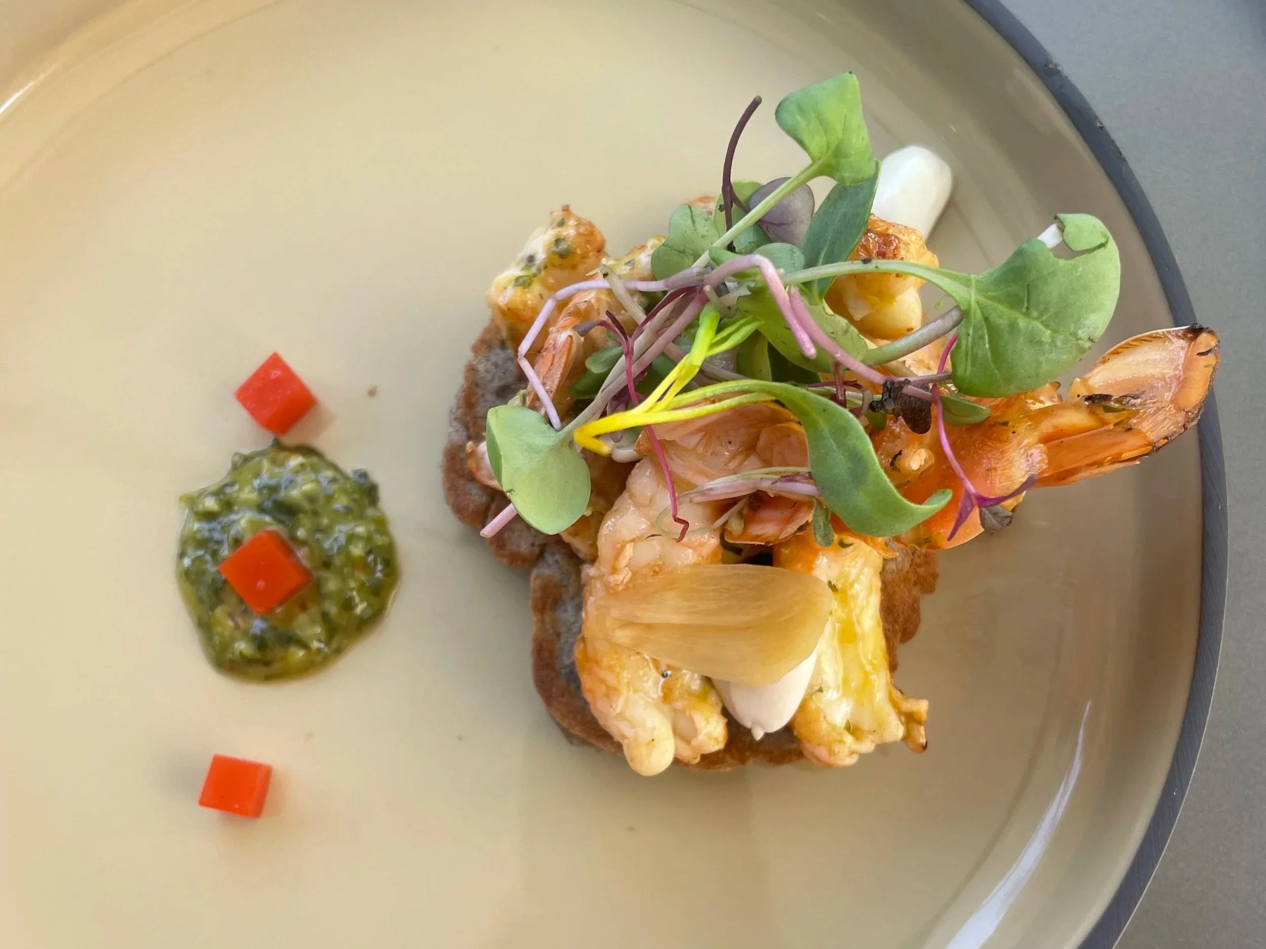 A plated dish with sautéed shrimp, microgreens, and a side of green sauce with diced red peppers on a pale yellow plate.