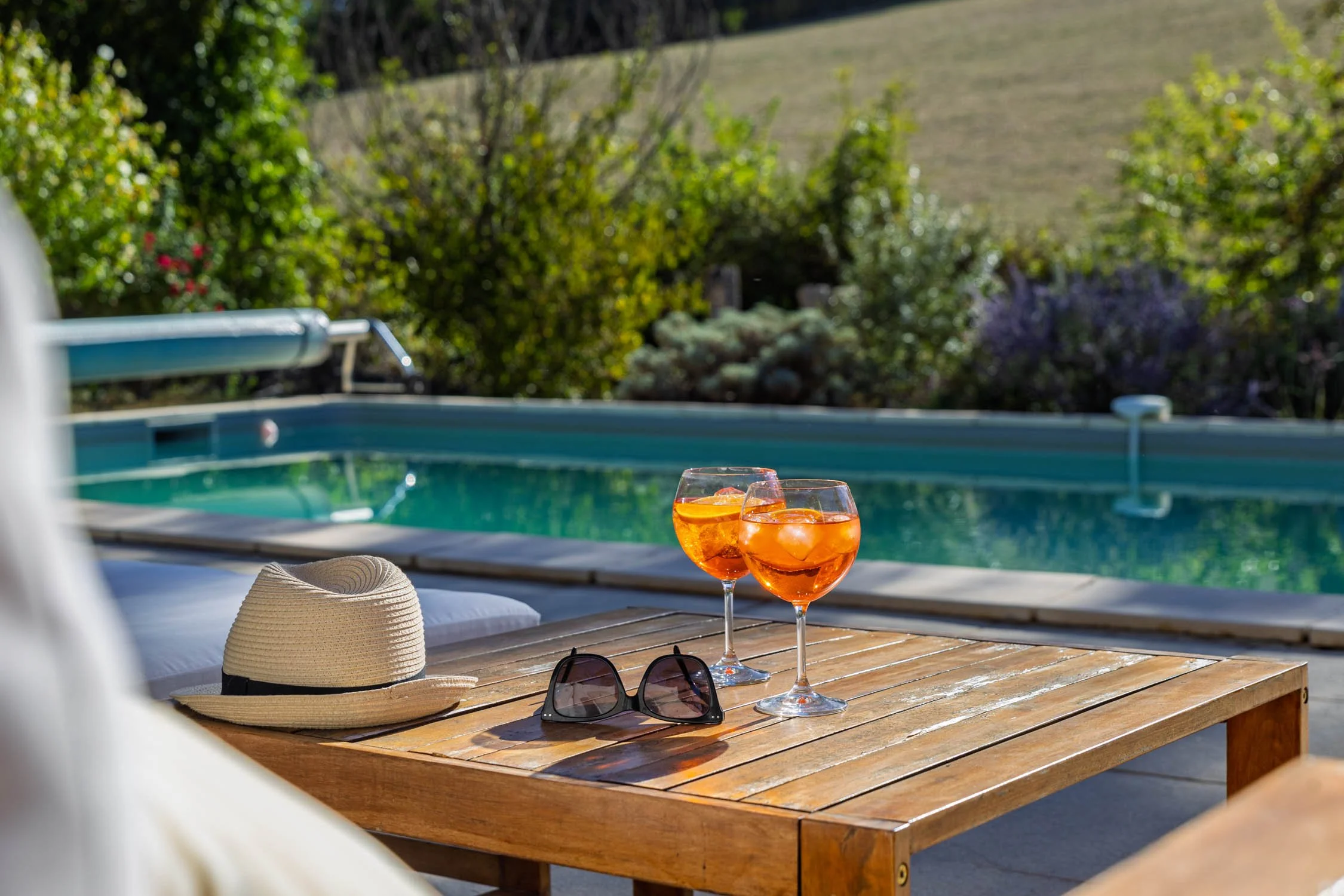 Two glasses of orange-colored drinks with ice cubes on a wooden table near a swimming pool, with a sunhat, a pair of sunglasses, and a towel also on the table. In the background, there are bushes and a grassy hillside.