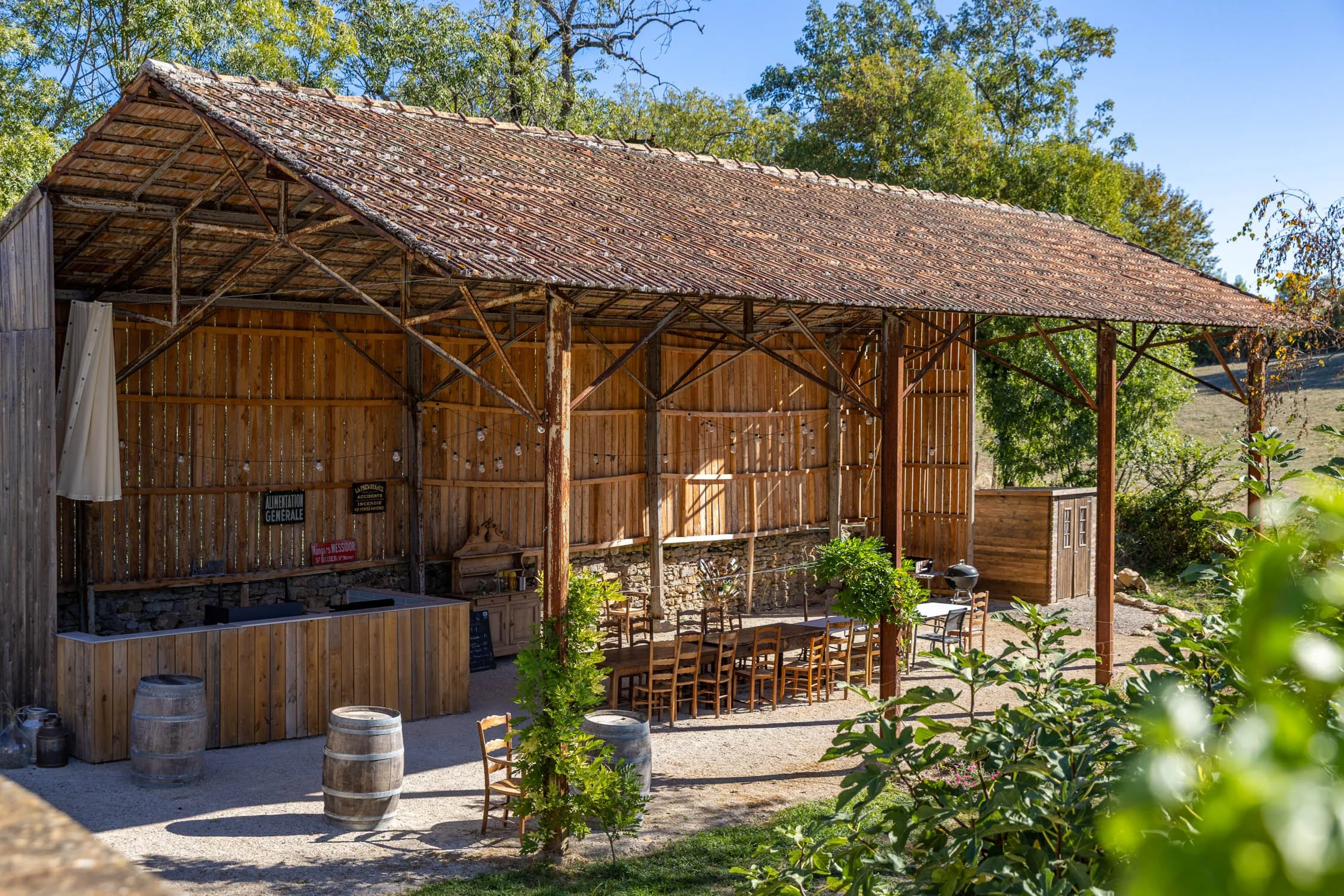 Outdoor rustic wooden structure with a tile roof, surrounded by trees, with signs, barrels, and tables and chairs for dining.