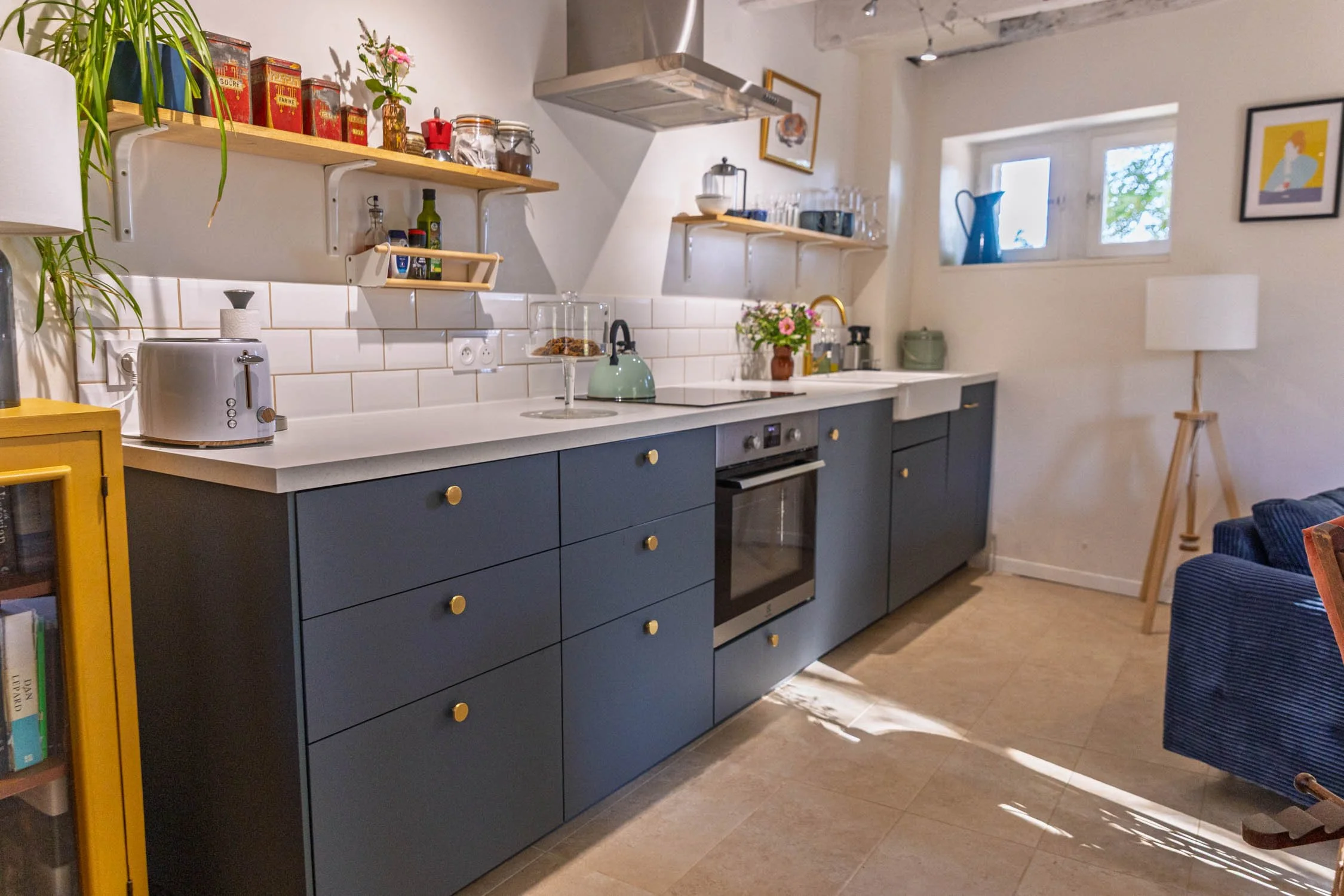 A modern kitchen with blue cabinets, white countertops, and a white subway tile backsplash. Open wooden shelves hold jars, bottles, and decorative items. There is a stainless steel oven, a green kettle, and a vase with pink flowers on the counter. The window lets in natural light and there is a floor lamp and framed artwork on the walls.