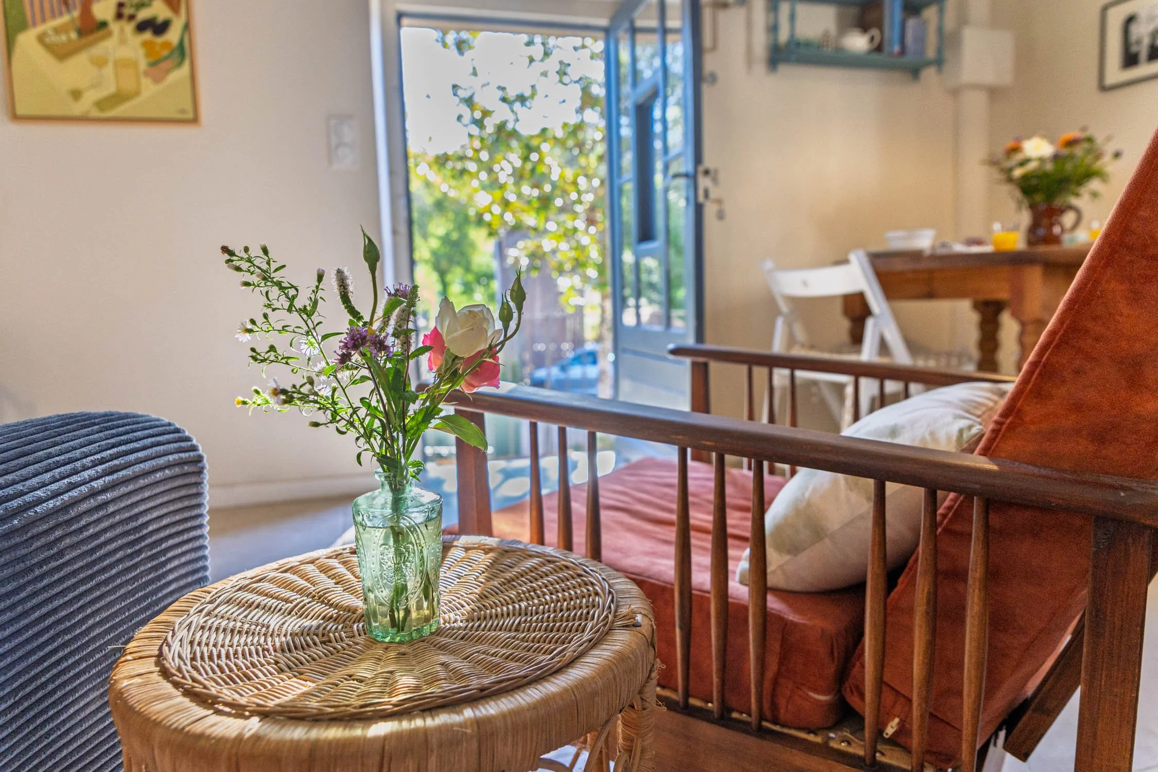 A cozy room filled with natural light from glass doors opening to a garden, with a small round wicker table with a green vase holding fresh flowers in the foreground, and a wooden dining table with a flower arrangement in the background.