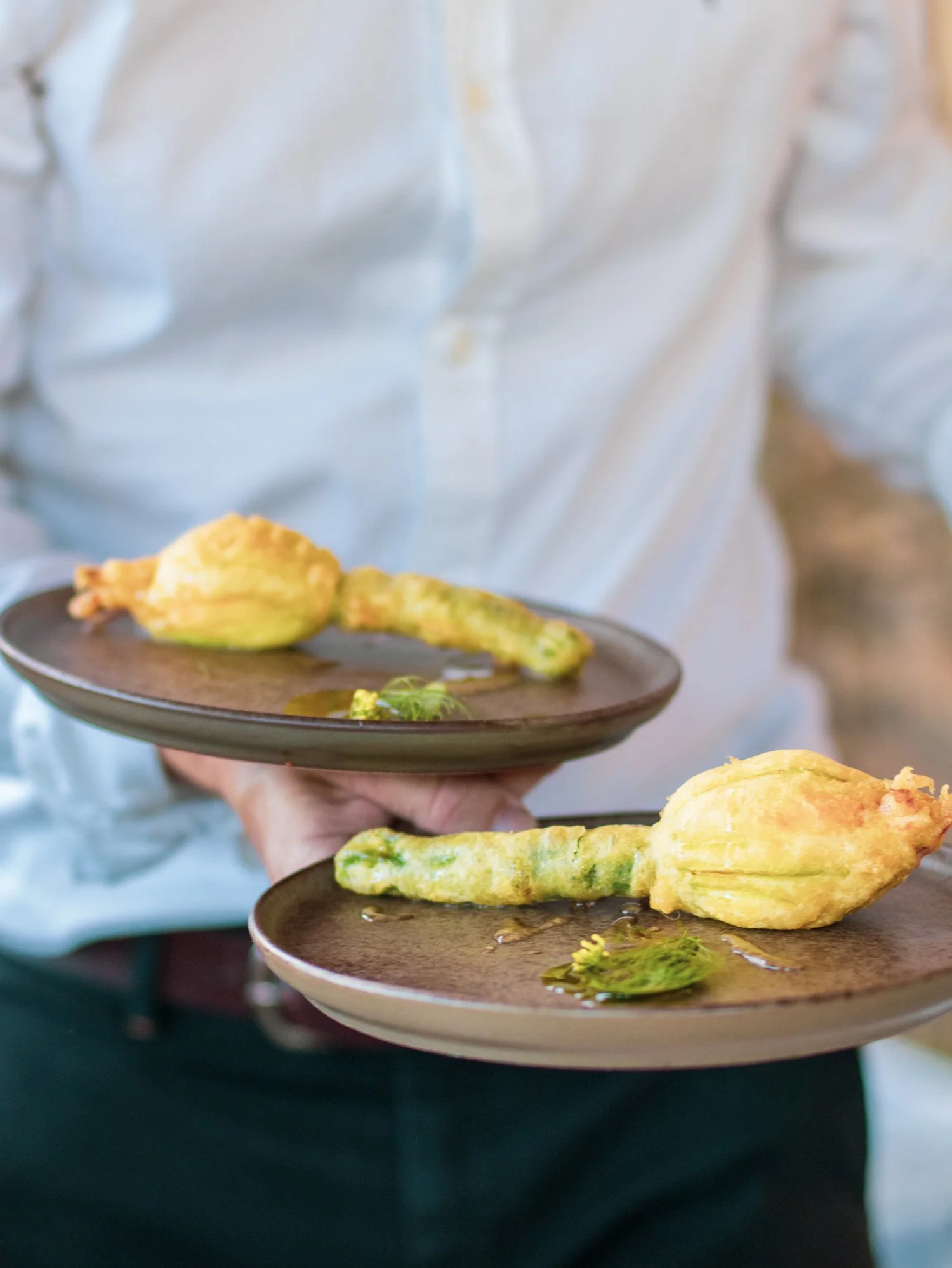 Two plates with fried courgette flowers and herbs held by a person in a white shirt.