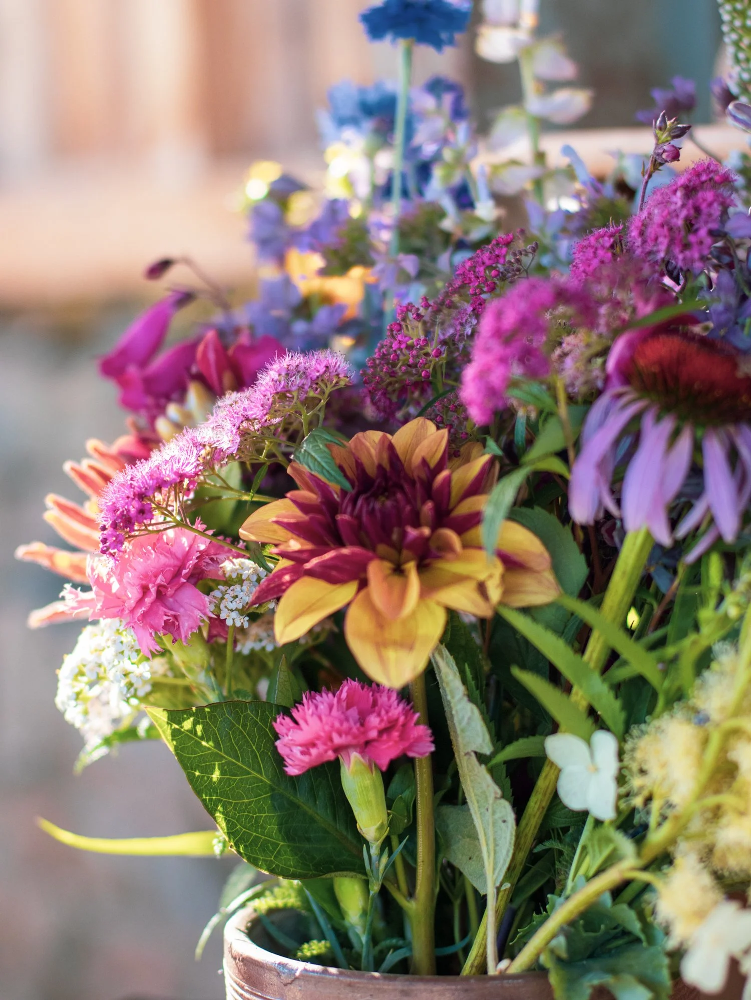 A diverse bouquet of colorful flowers in a rustic clay pot, including pink, purple, yellow, and blue blooms, with lush green leaves.