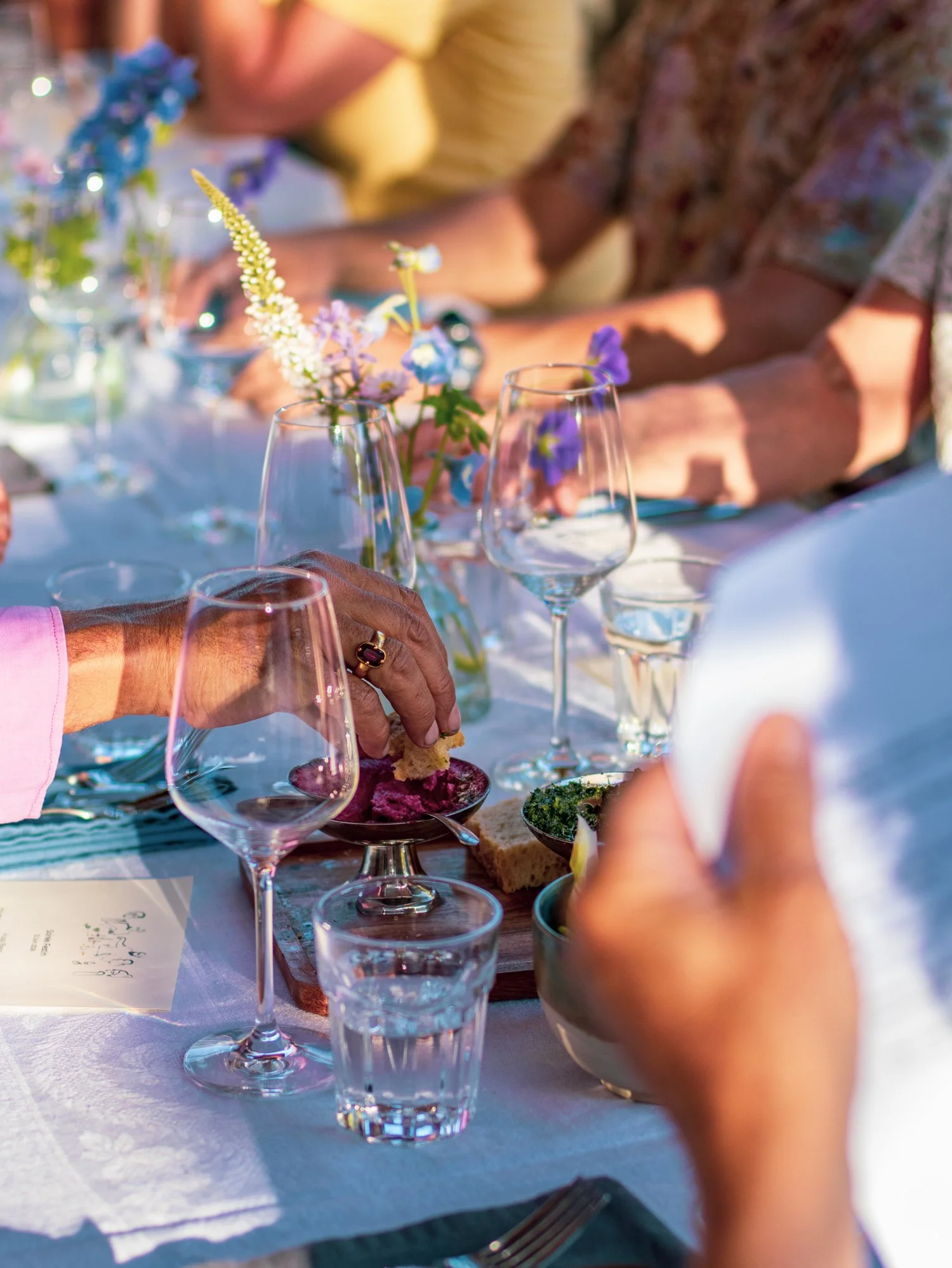Table set with wine glasses, flowers in small vases, and plates of food, with people in the background