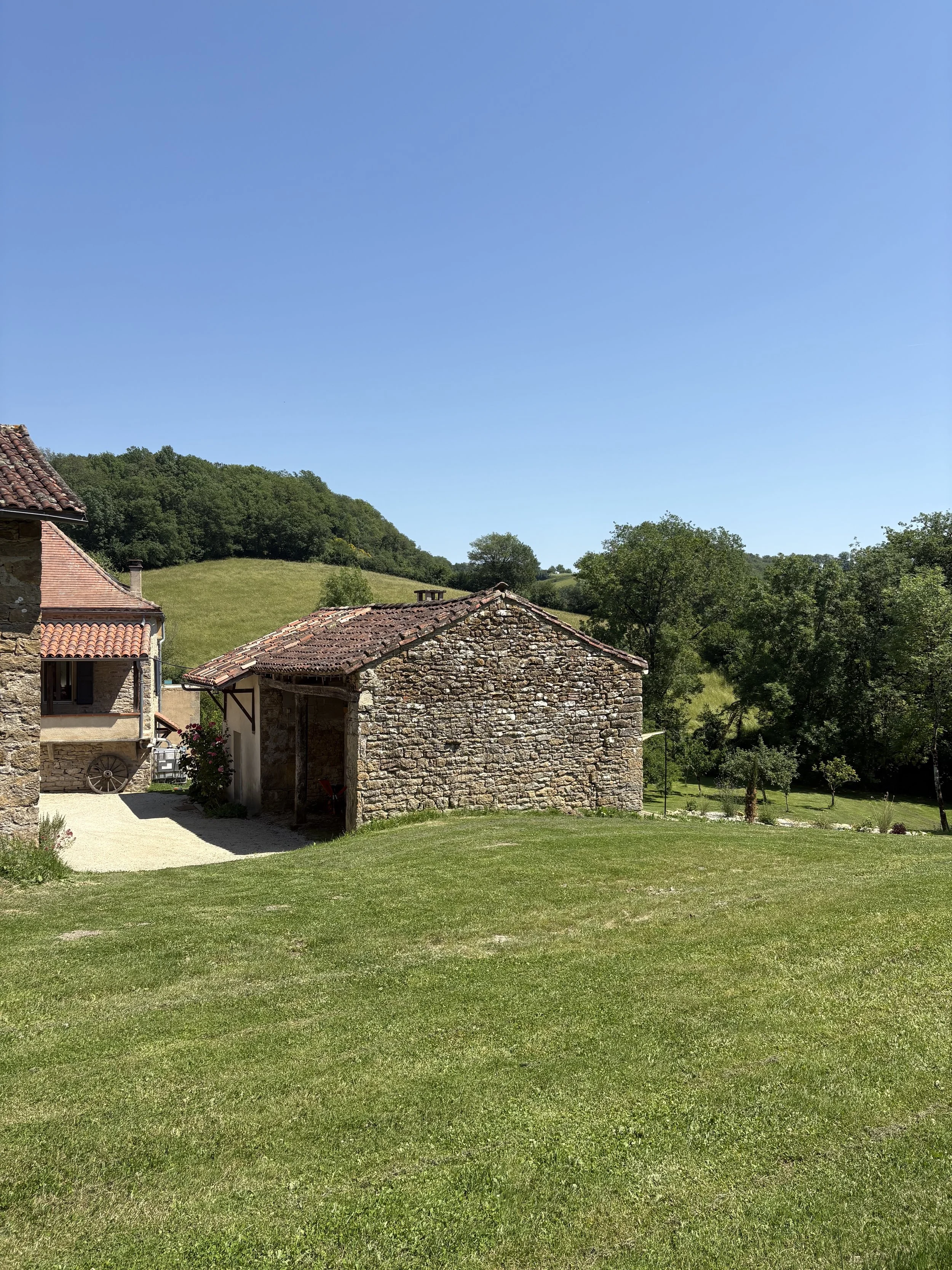 A scenic image of green grassy field with rustic stone and brick buildings in the foreground and a line of trees and a hill in the background under a clear blue sky.