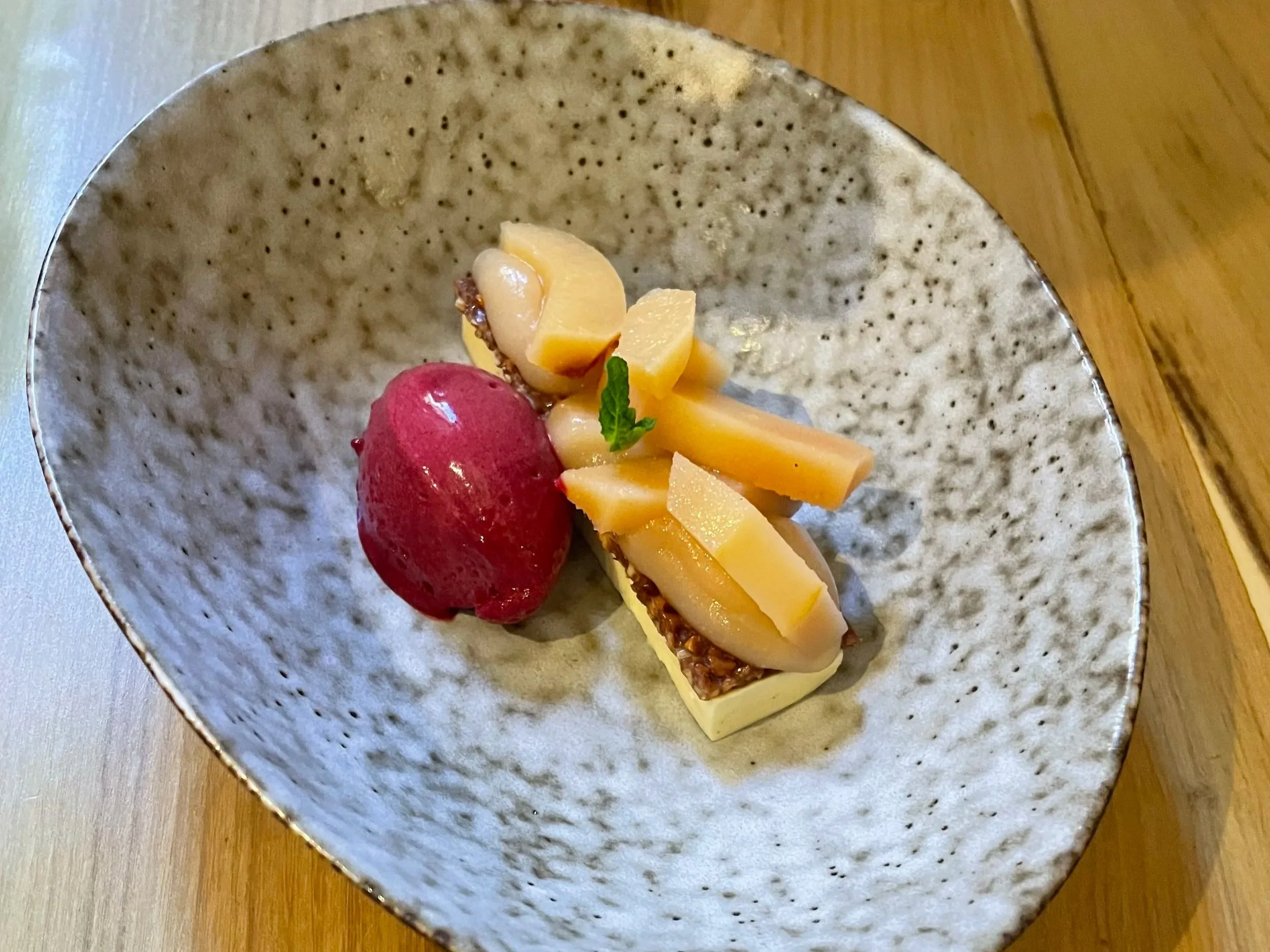 A ceramic bowl with a speckled glaze contains a dessert with sliced peaches, a scoop of raspberry sorbet, and a sprig of mint.