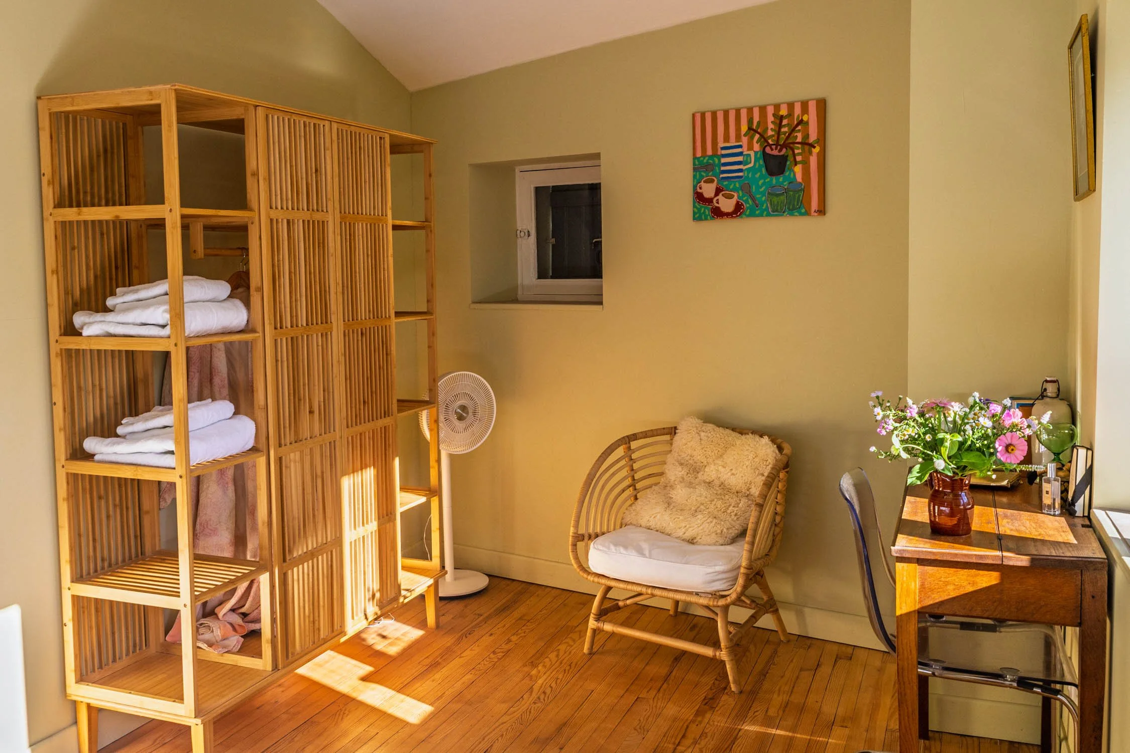 A cozy living room with wooden flooring, a wooden shelving unit with folded towels, a wicker chair with a fluffy cushion, a small fan, a wooden table with a vase of pink and purple flowers, a painting on the wall, and a small window.