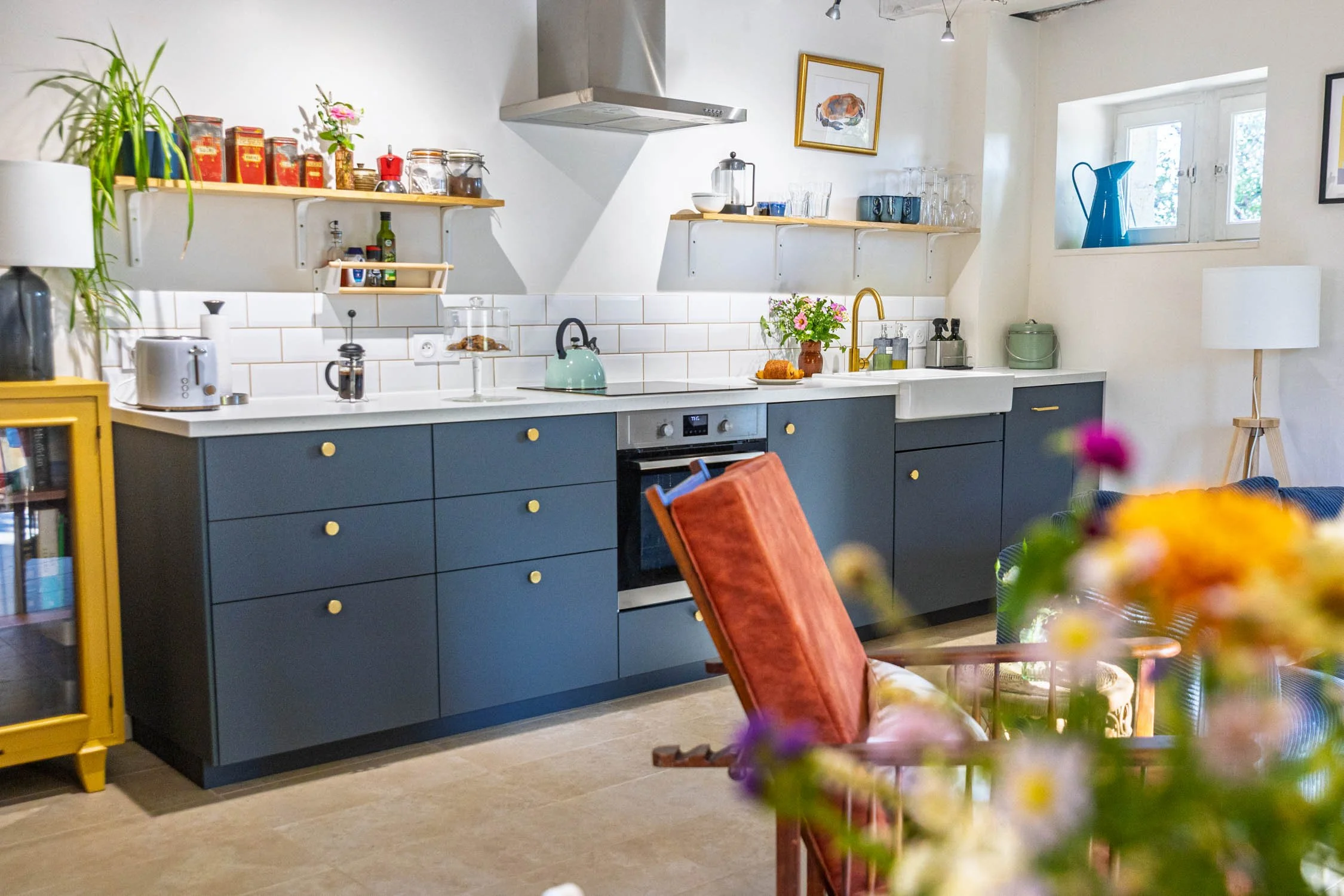 A modern kitchen with light-colored walls, a white tiled backsplash, and dark blue cabinets with yellow knobs. There are open shelves with jars, cans, glasses, and a framed picture on the wall. A window with blue water pitcher on the sill and a potted plant on the left. An orange chair with a pink cushion is in the foreground, and a table with a floral centerpiece is partially visible.