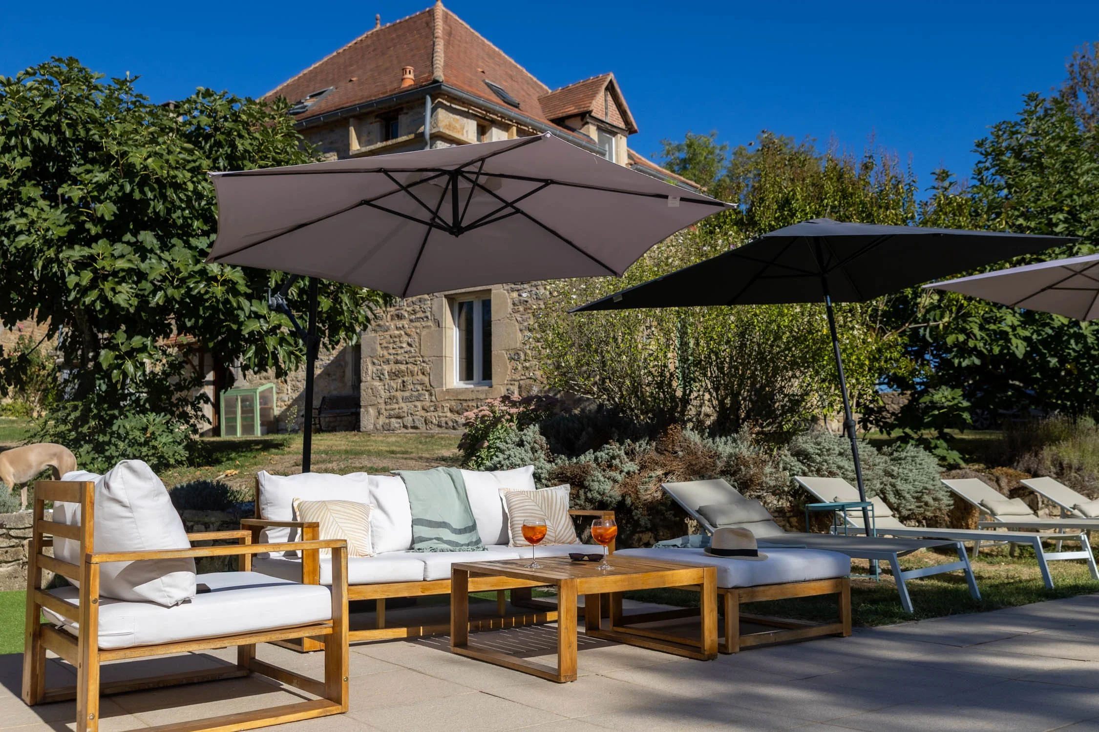 Backyard patio scene with wooden outdoor furniture, white cushions, and glass of rosé on the table, with large umbrellas, green trees, and a stone house in the background under a clear blue sky.